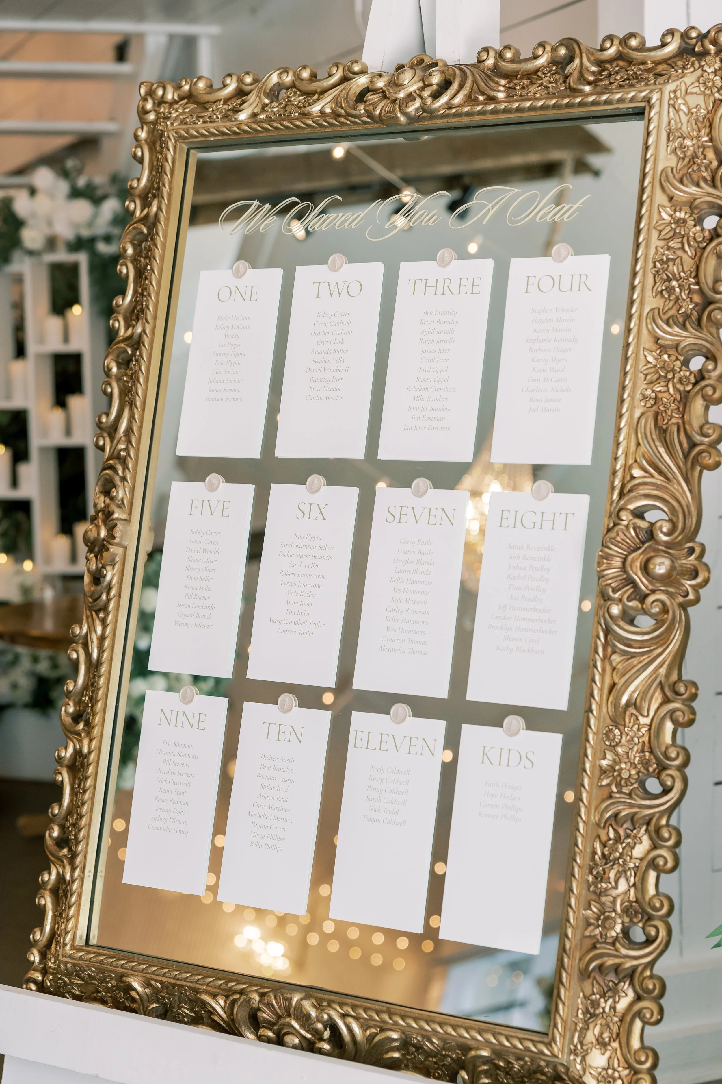 A gold ornate framed mirror with a seating chart for a wedding, showing table numbers one through eleven and kids, with guests' names listed under each table number.