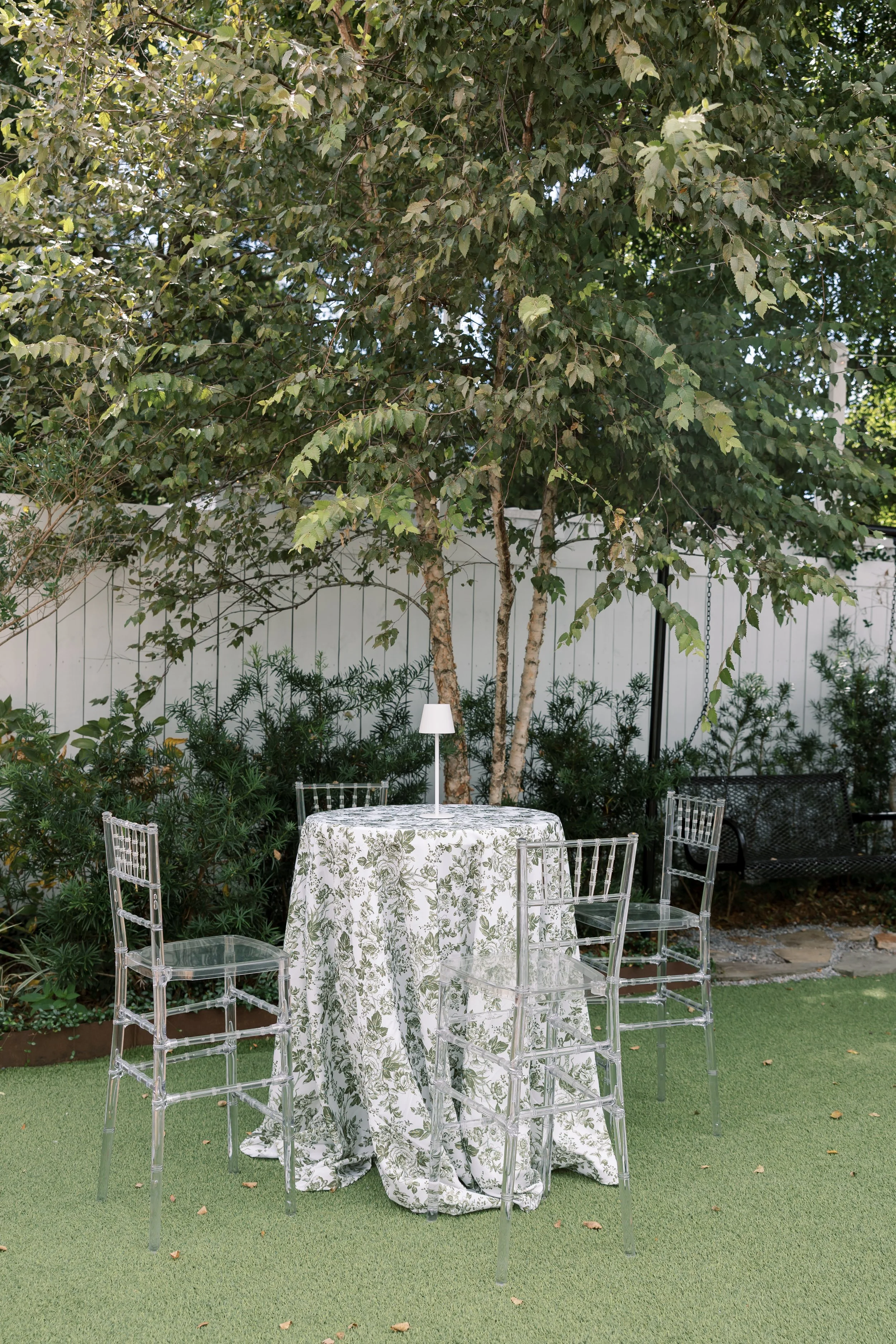 Outdoor patio setup with a round table covered with a white and green floral tablecloth, surrounded by four clear acrylic chairs, a small lamp on the table, and greenery including trees and bushes in the background.