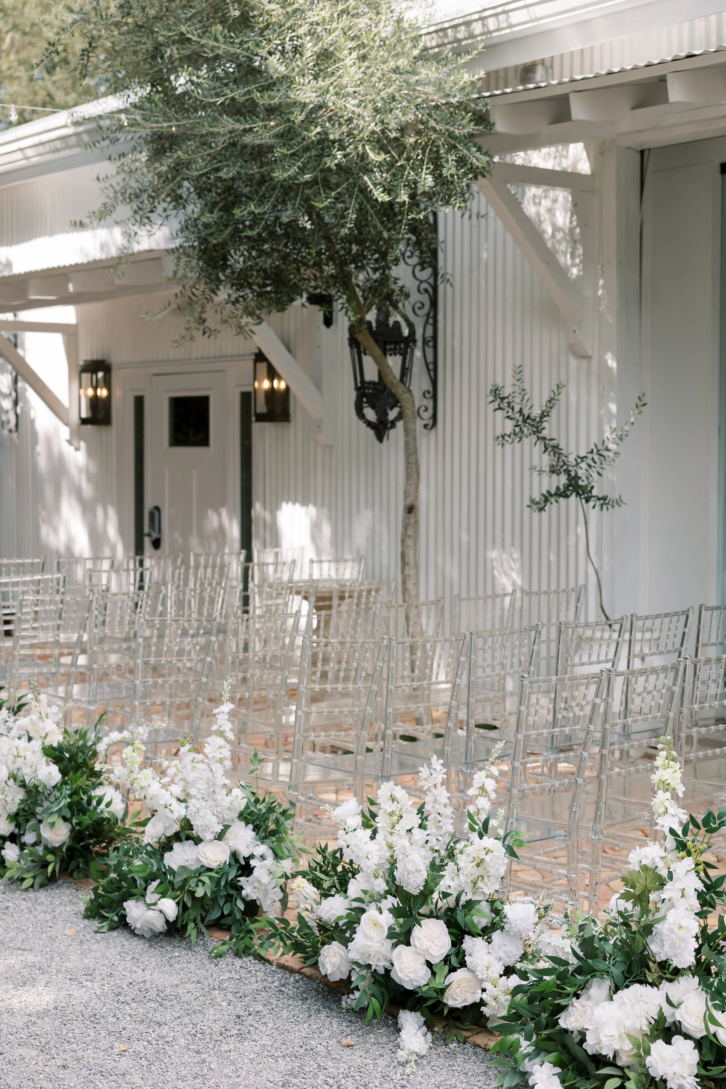 Outdoor wedding ceremony setup with clear chairs and white floral arrangements along the aisle, against a white building with greenery and decorative black lanterns.