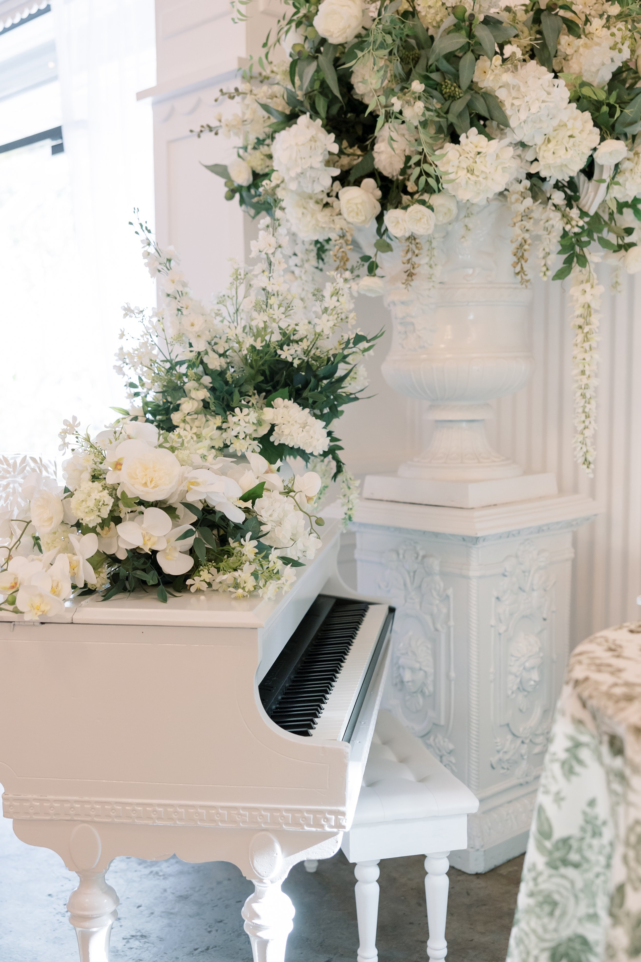 White grand piano decorated with white flowers and greenery, featuring large floral arrangements and a white upholstered bench, in a bright, elegant room.