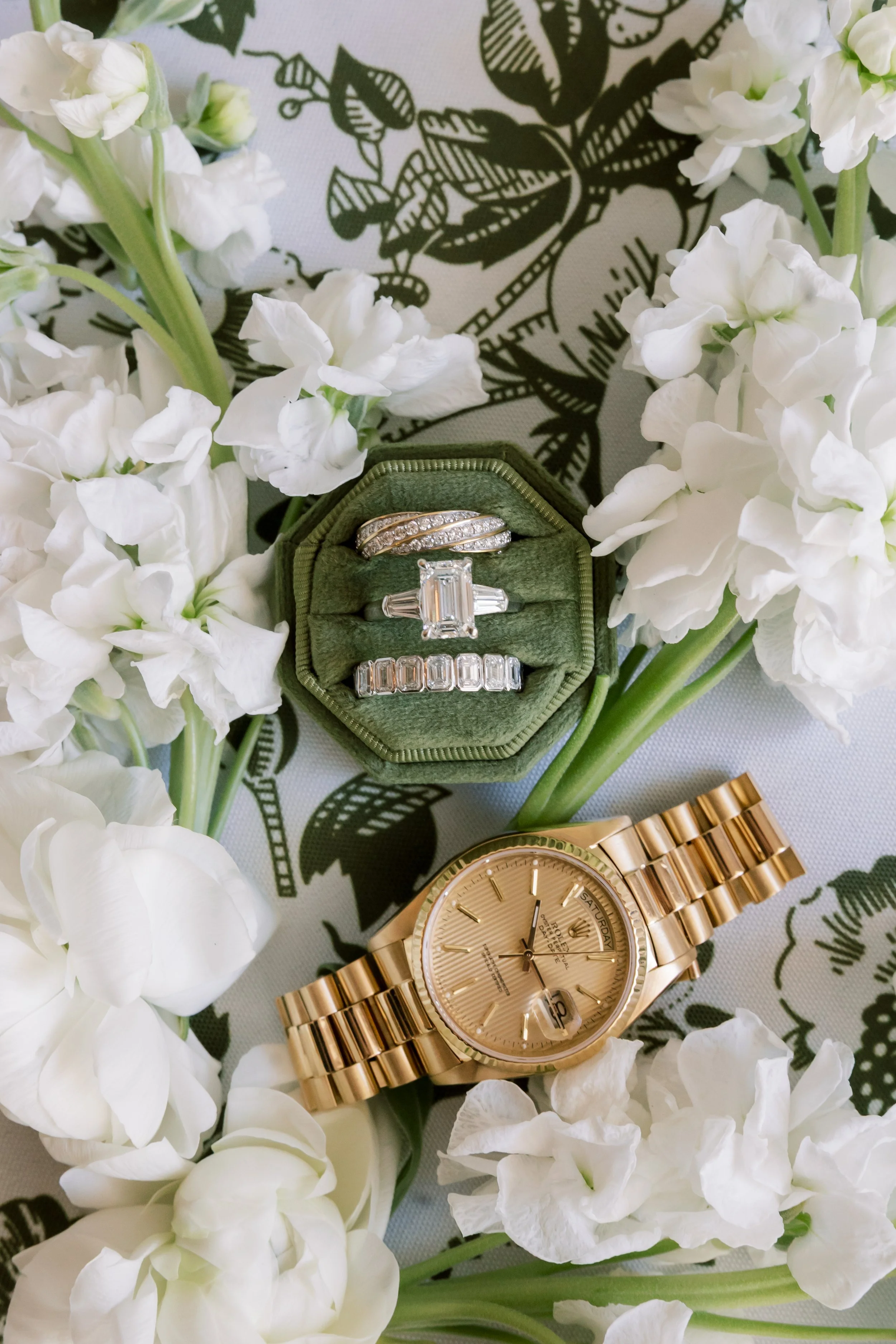 A gold Rolex watch, a green jewelry box with diamond rings, and white flowers surrounding the items.