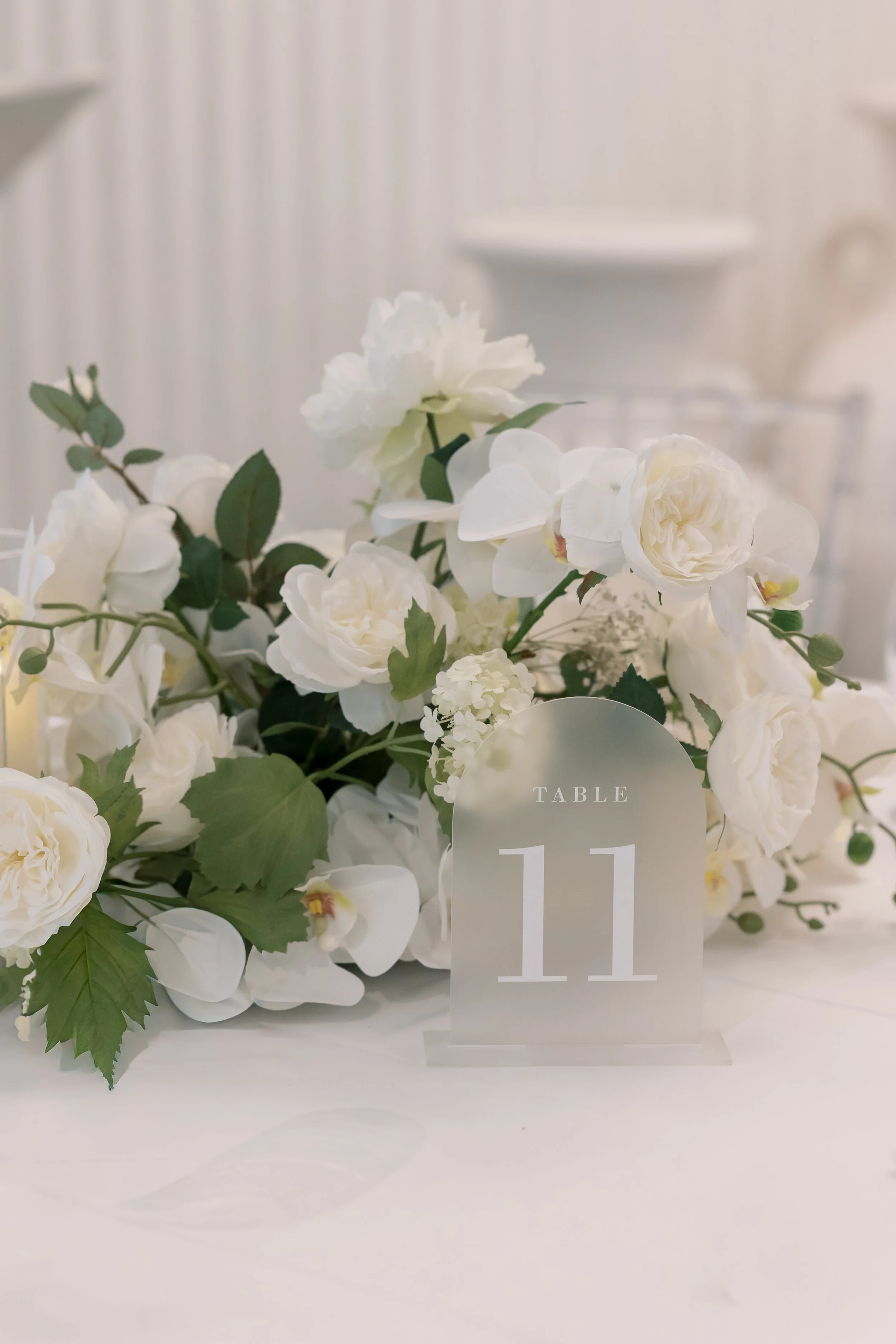 White floral centerpiece on table with a table number sign displaying 'Table 11' in an elegant font, set in a white or neutral-colored venue.