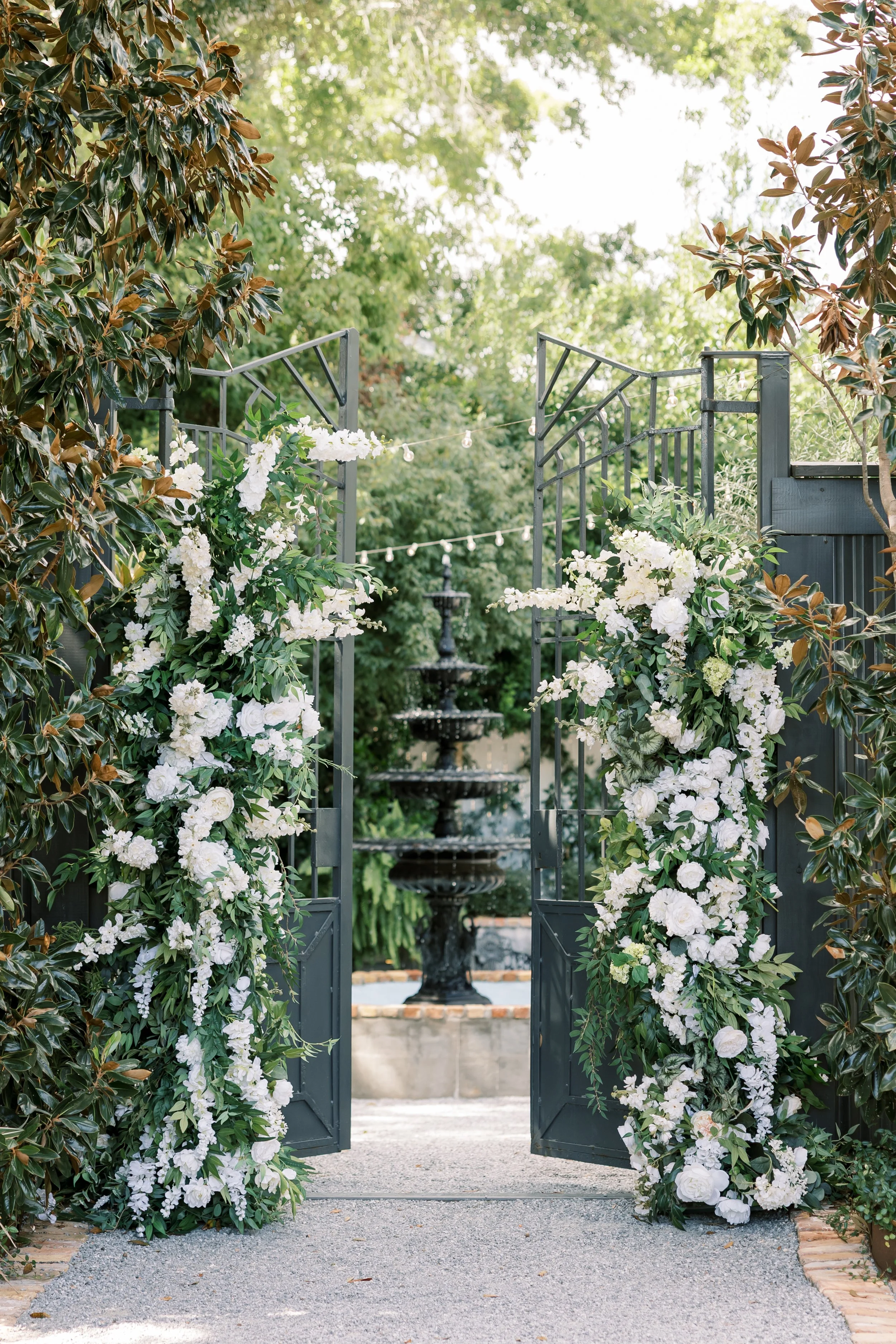 Open black metal gate decorated with white floral arrangements and lush green foliage, leading to a fountain in a garden.