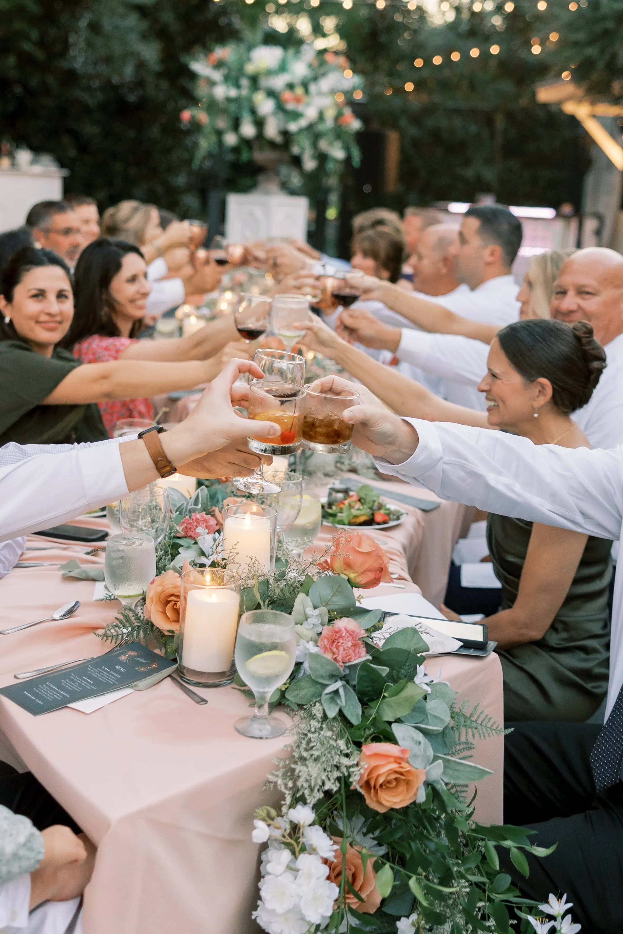 People gathered at a long table celebrating, raising their glasses in a toast, with candles and floral decorations on the table.