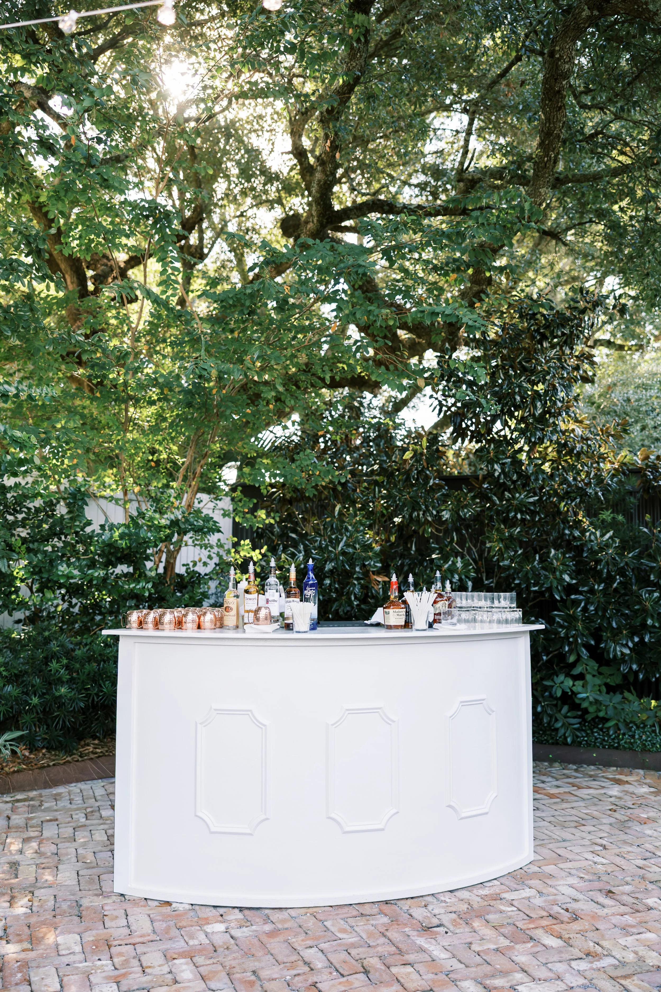 Outdoor bar with various bottles and glasses, set on a white curved bar counter on a brick patio, surrounded by lush green trees.