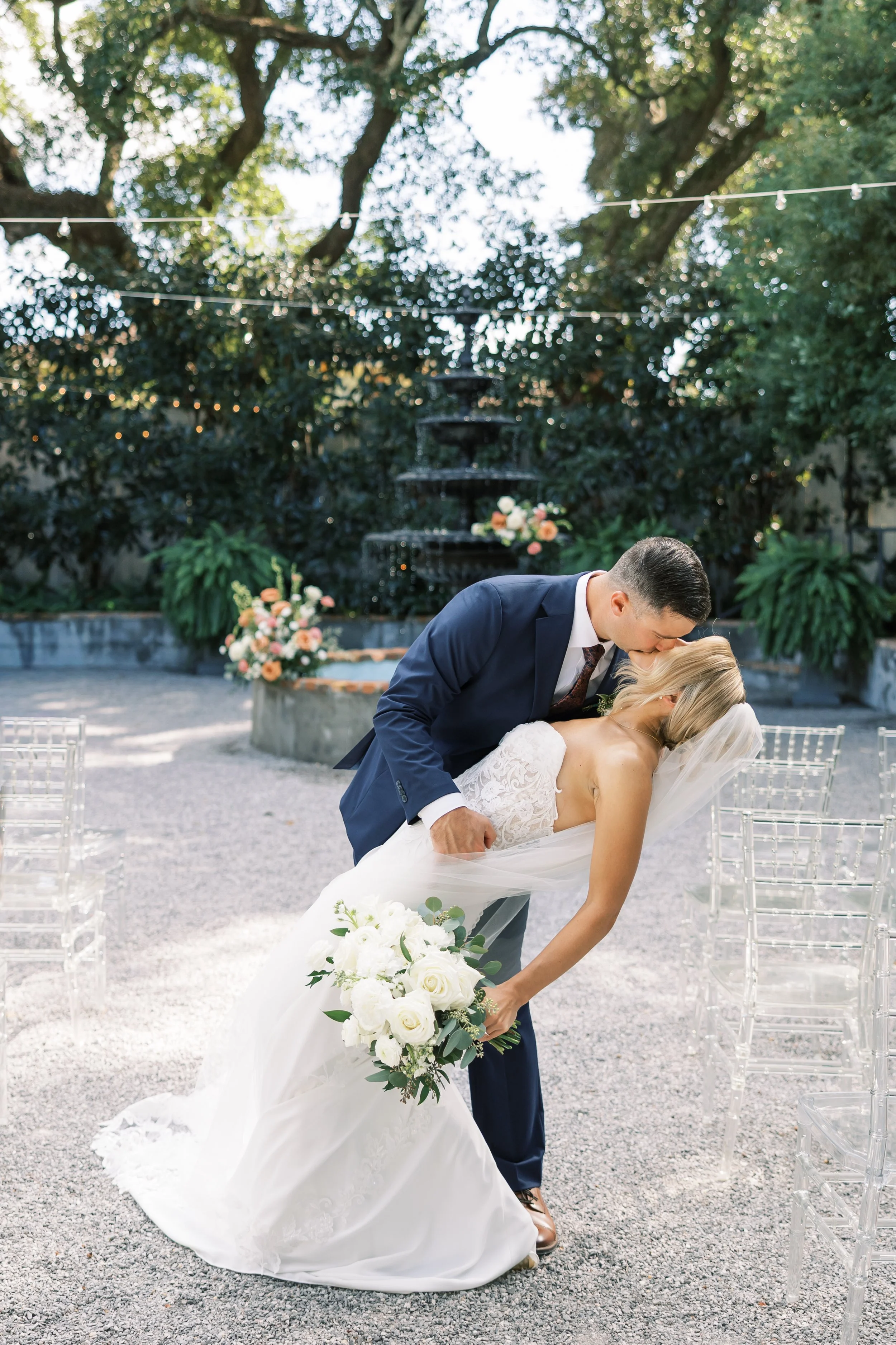 A newlywed couple sharing a kiss outdoors at their wedding venue, with the groom dipping the bride who is holding a bouquet of white roses.