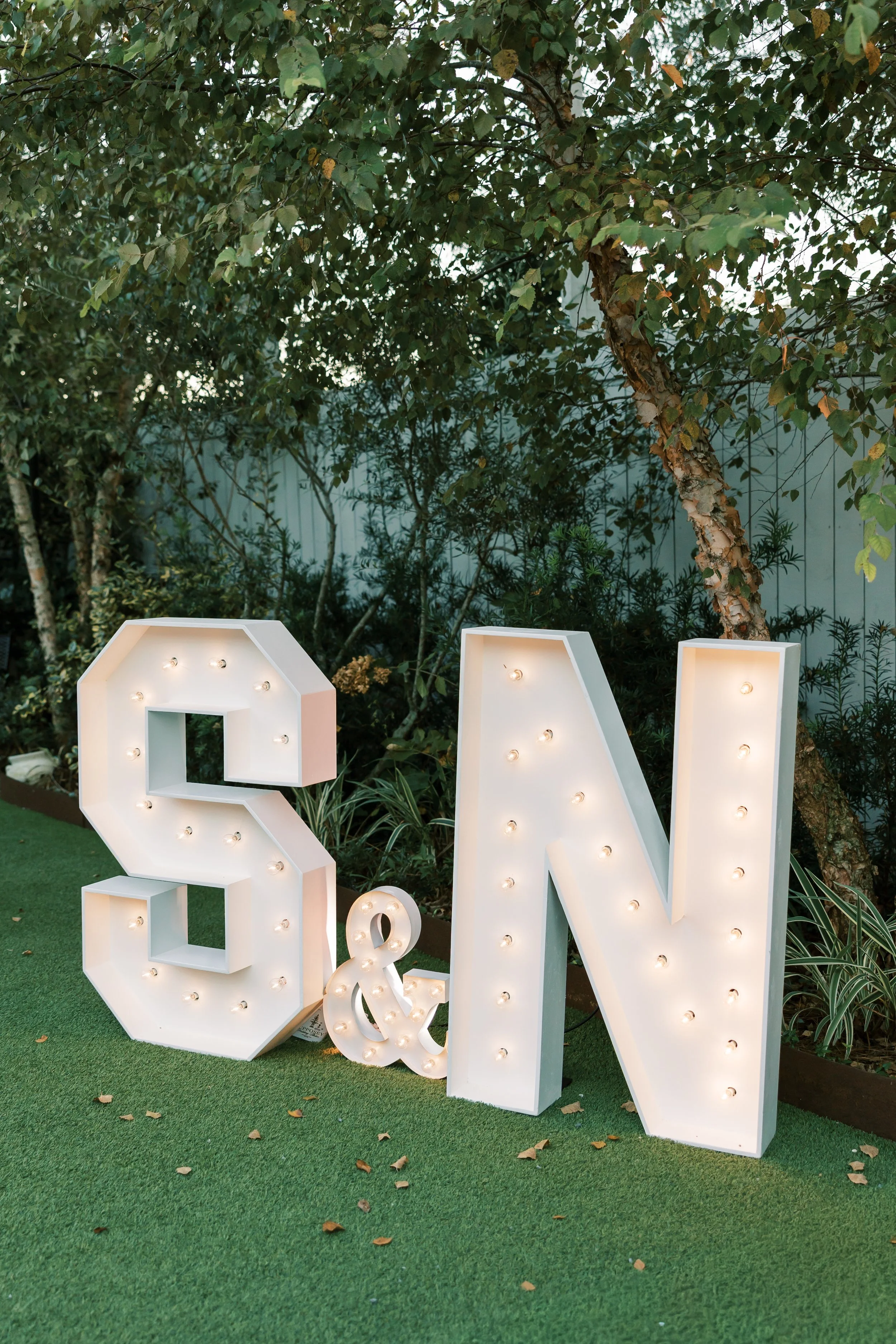 Decorative marquee lights spelling out the letters 'S', '&', and 'N' on a grassy outdoor area with trees and plants in the background of a Pensacola Wedding Venue.