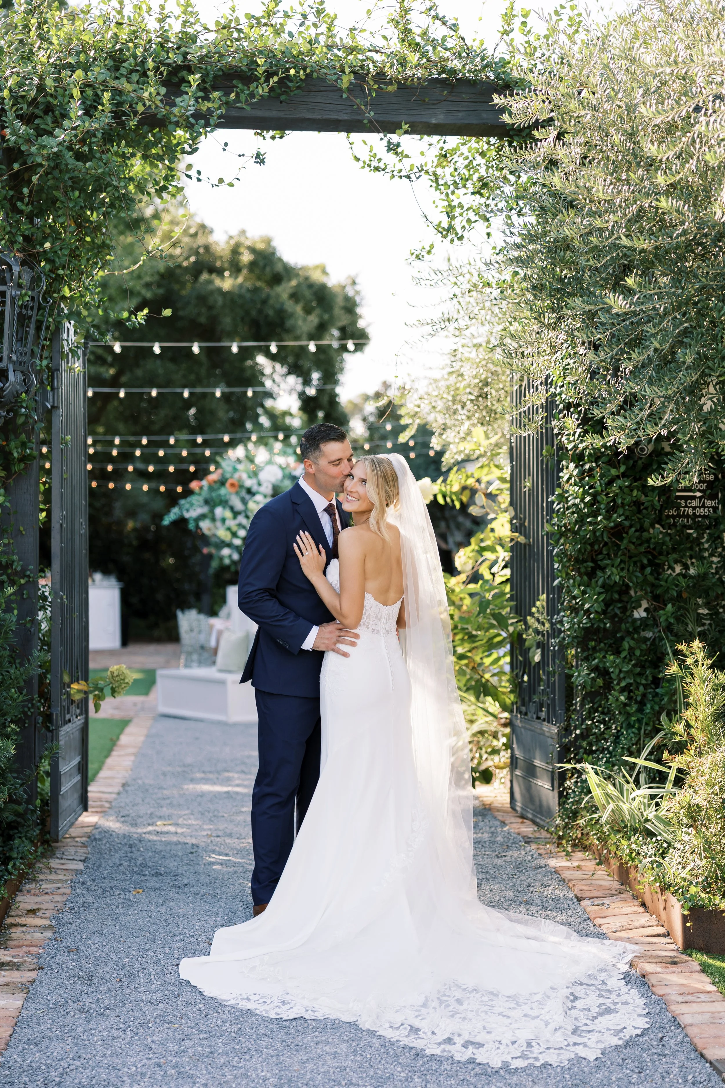 A newlywed couple shares a kiss during their wedding photos at an outdoor garden venue, with string lights hanging overhead and greenery surrounding them at a Pensacola Wedding Venue.