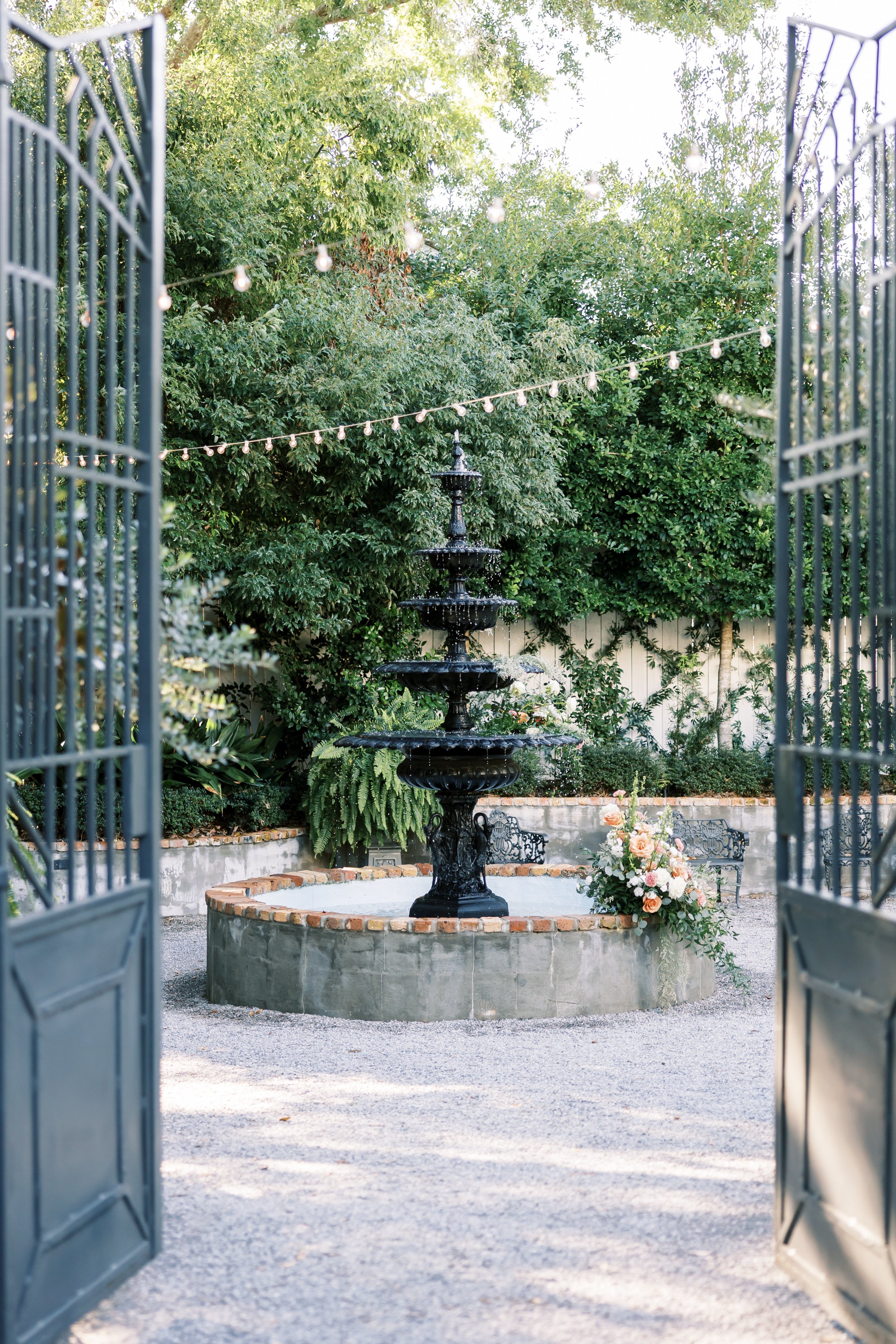View through open gates of a garden fountain with multiple tiers, surrounded by greenery and flowers, with string lights overhead.