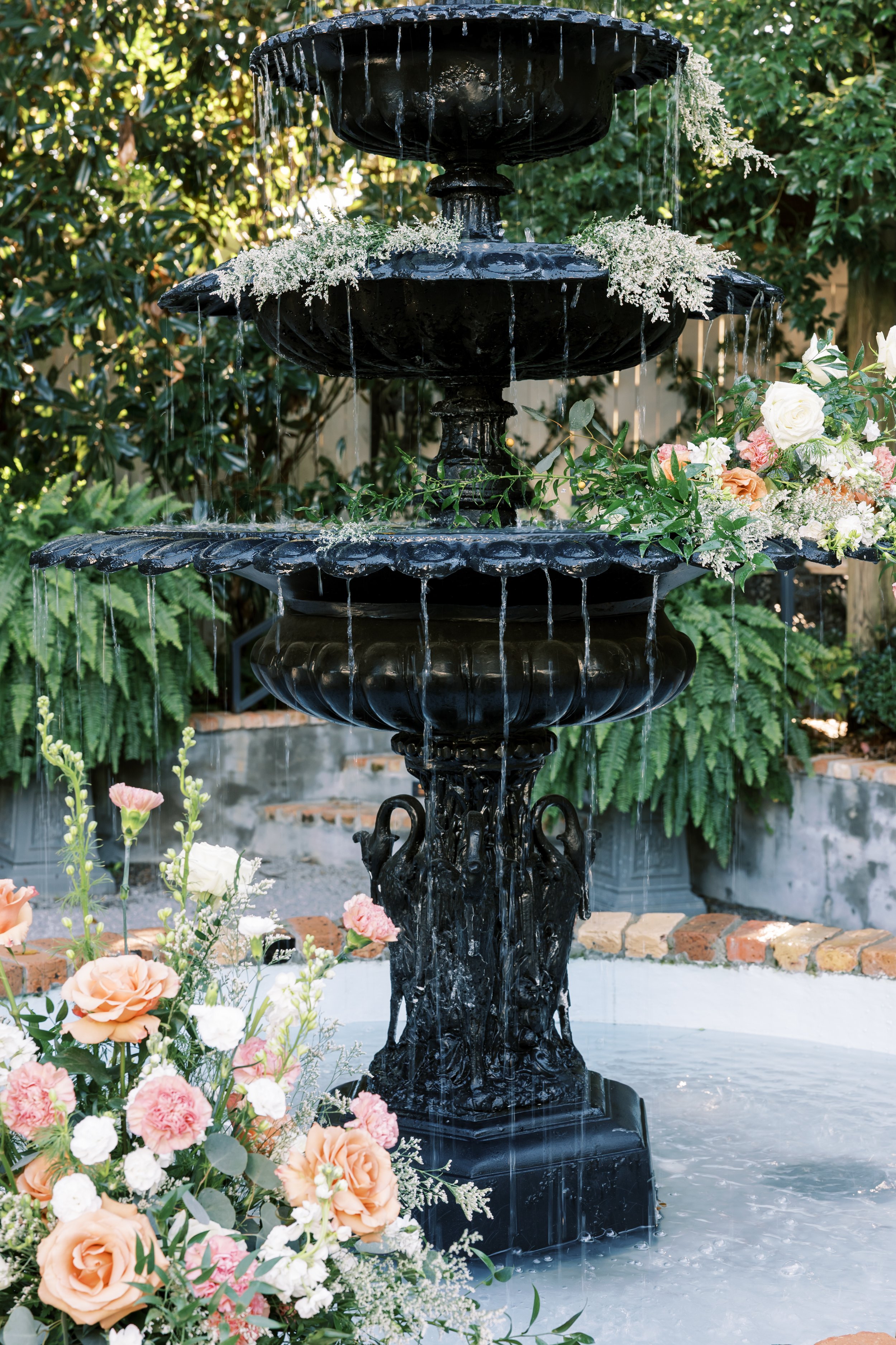 A black, three-tiered outdoor fountain with water flowing down, decorated with white flowers and surrounded by a garden with green foliage and pink and white flowers.
