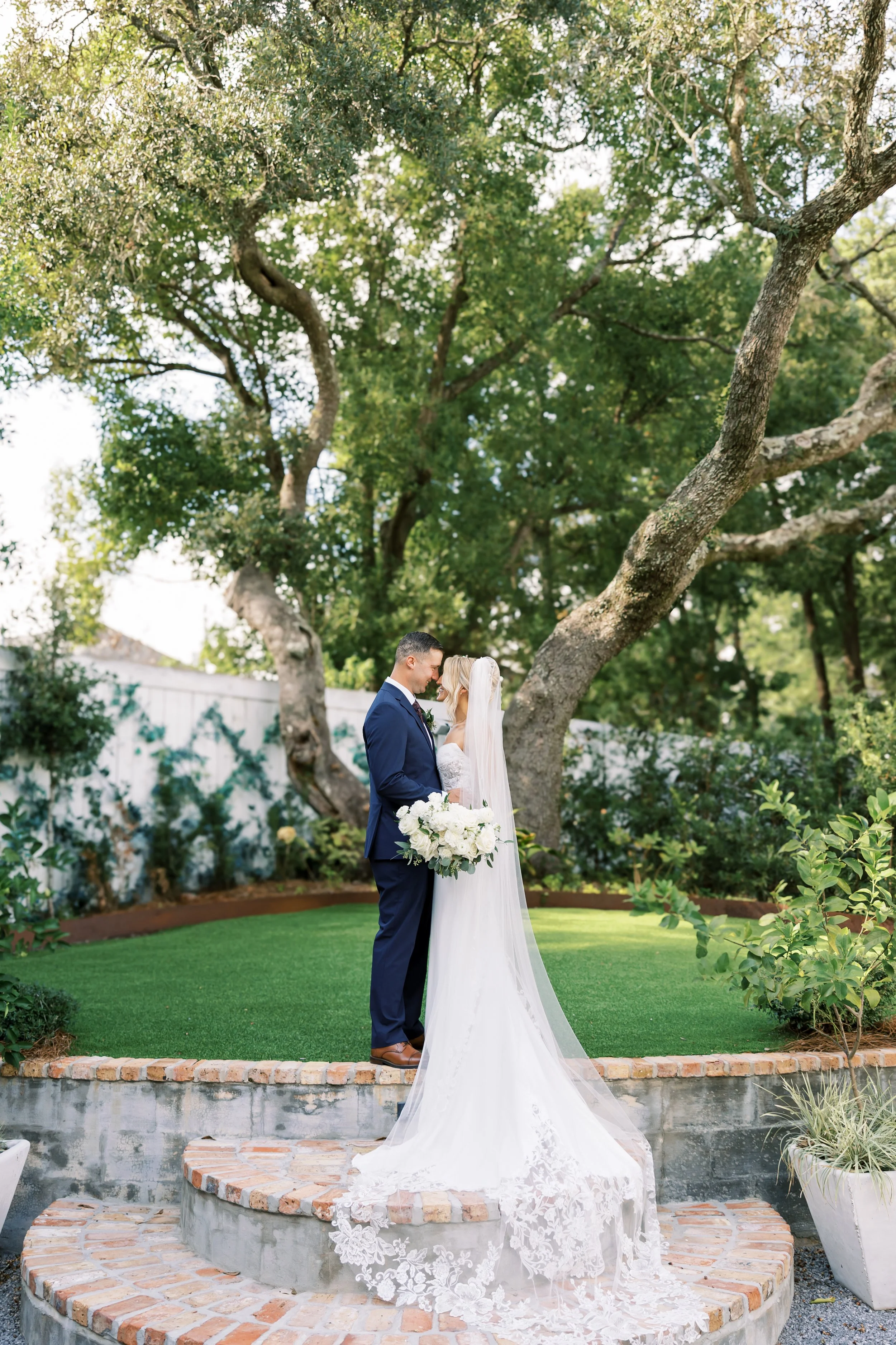 Bride and groom standing close together on a small brick and concrete platform in a garden with large trees and green shrubs, exchanging vows on a Pensacola sunny day.