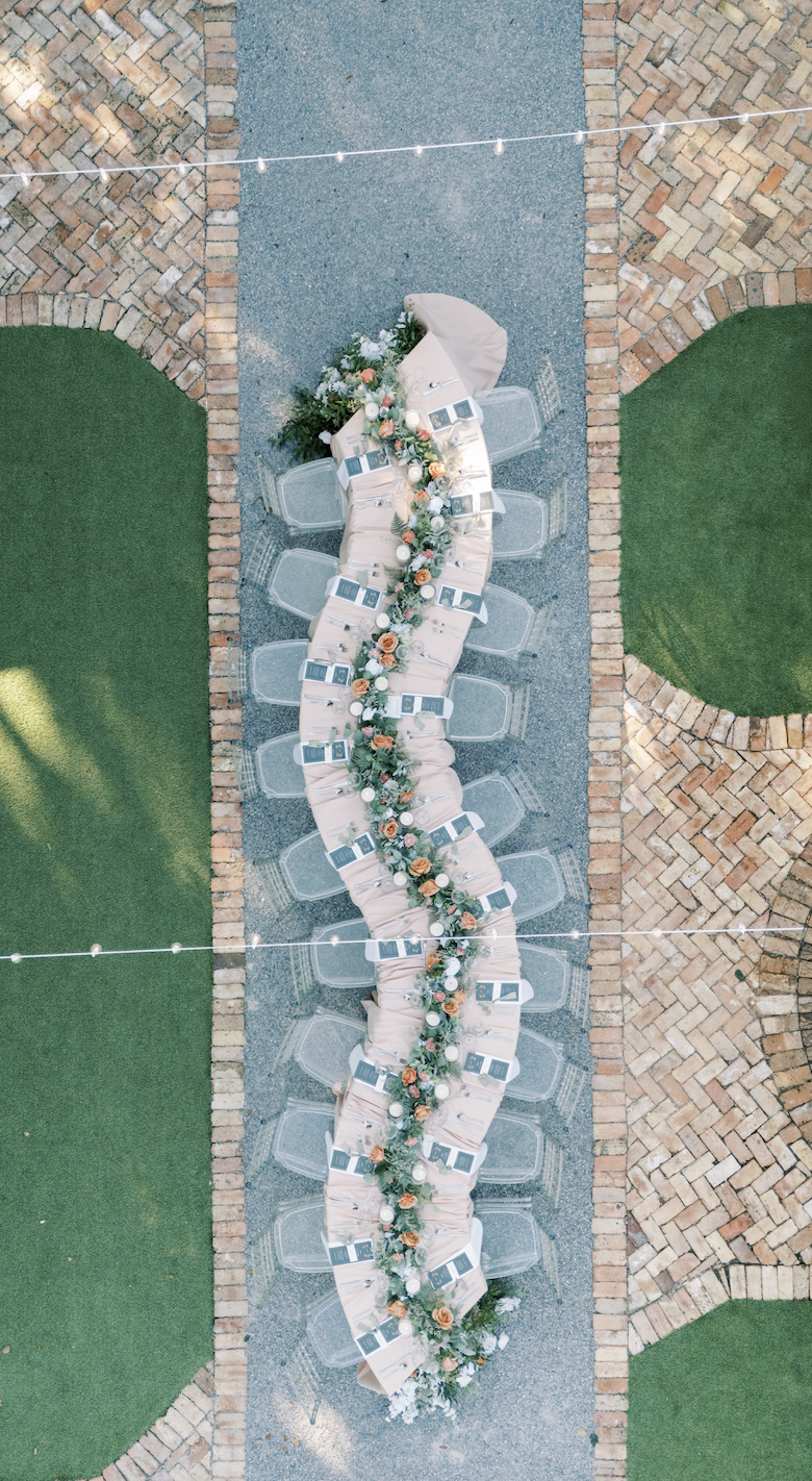 Overhead view of a long outdoor banquet table decorated with a floral garland, set with beige tablecloths, clear chairs, and place settings, on a gravel surface surrounded by brick and green grass areas with string lights overhead in Pensacola.