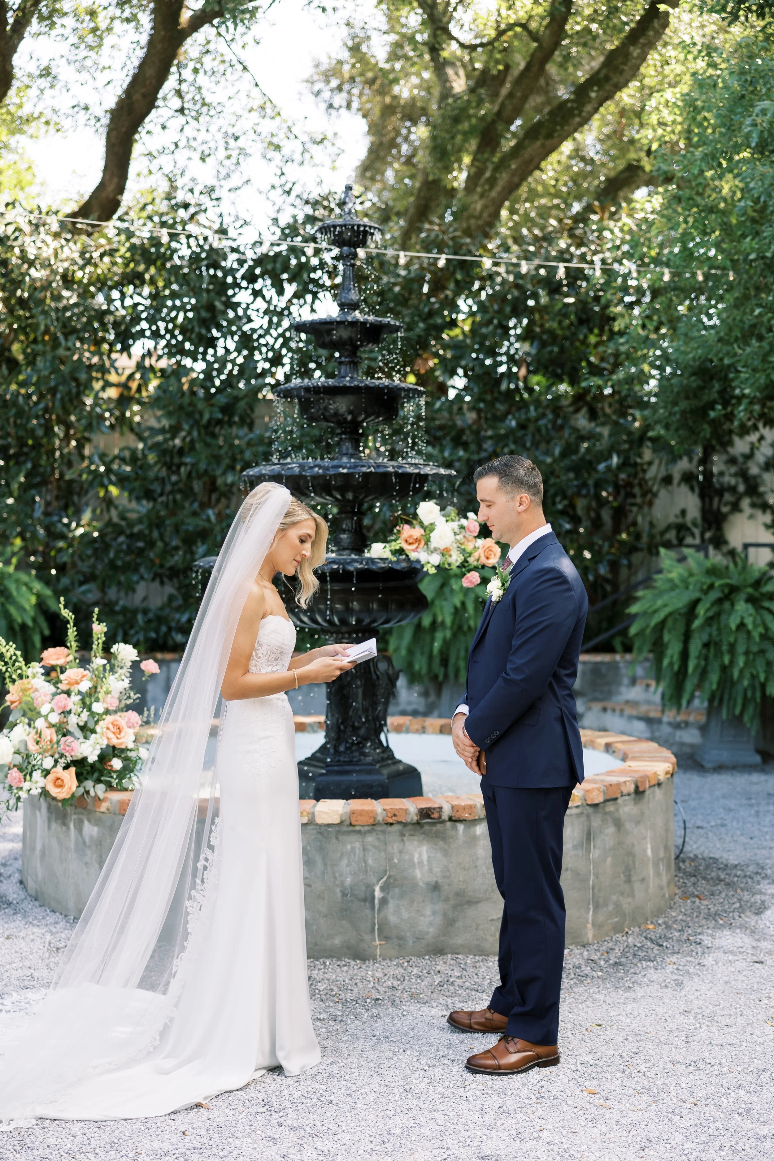 Bride and groom standing in front of a fountain, engaged in their wedding vows during an outdoor ceremony, with flowers and greenery surrounding them in Pensacola, Florida