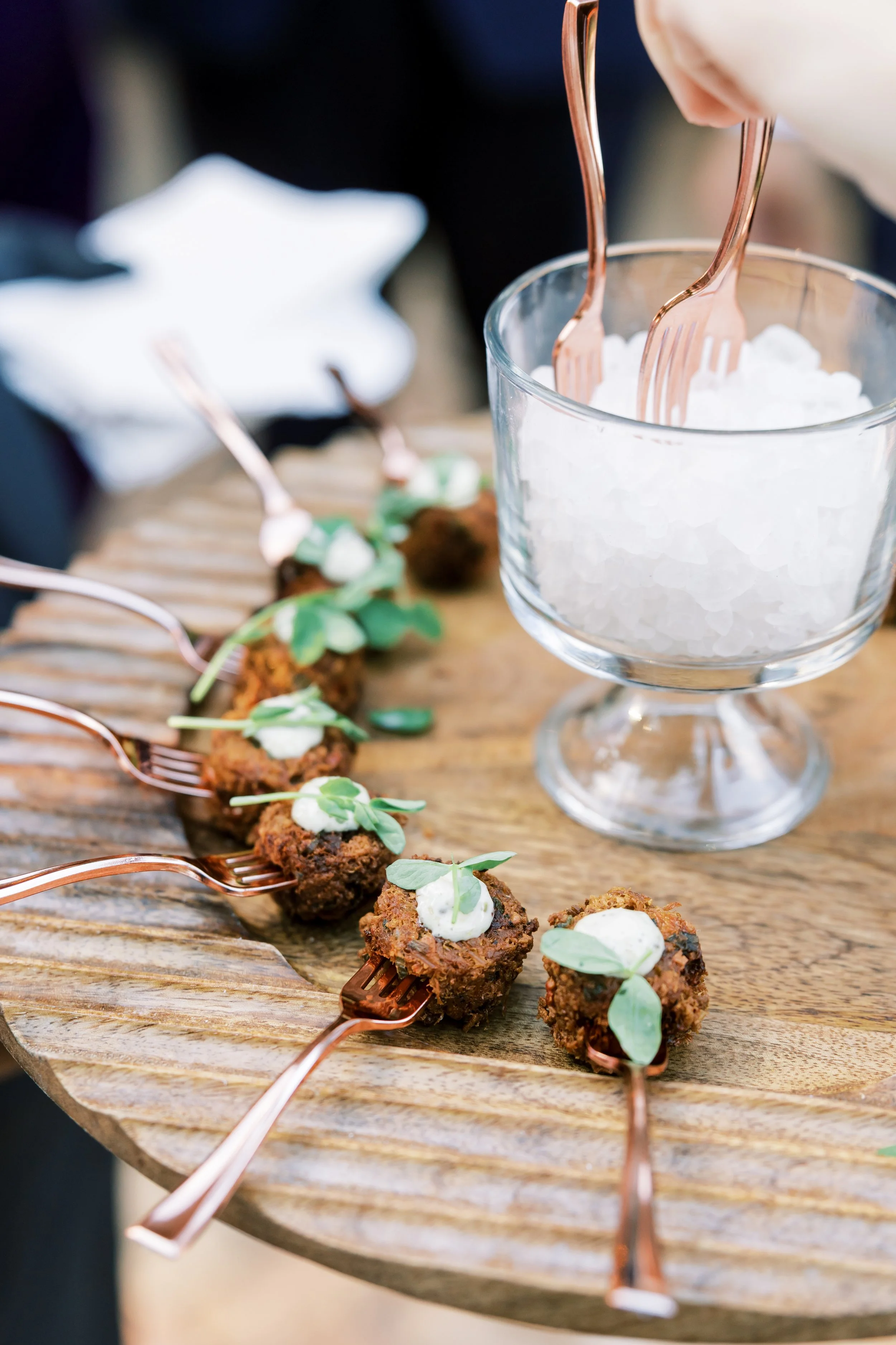 Skewers of fried food garnished with green herbs, placed on a rustic wooden serving tray, with a glass container of ice and forks.