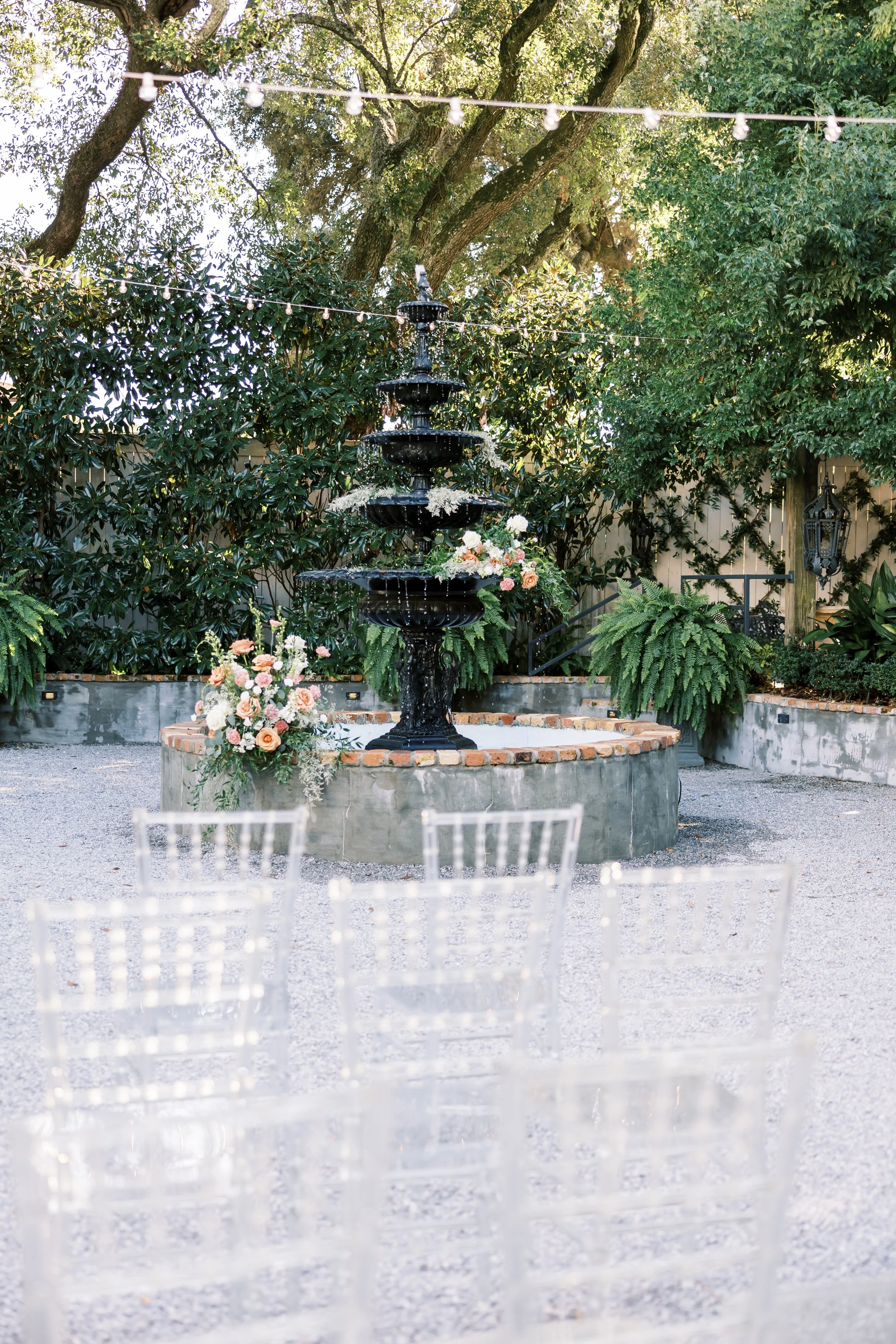 Outdoor event setup with a black fountain surrounded by greenery, floral arrangements, string lights, and white chairs.