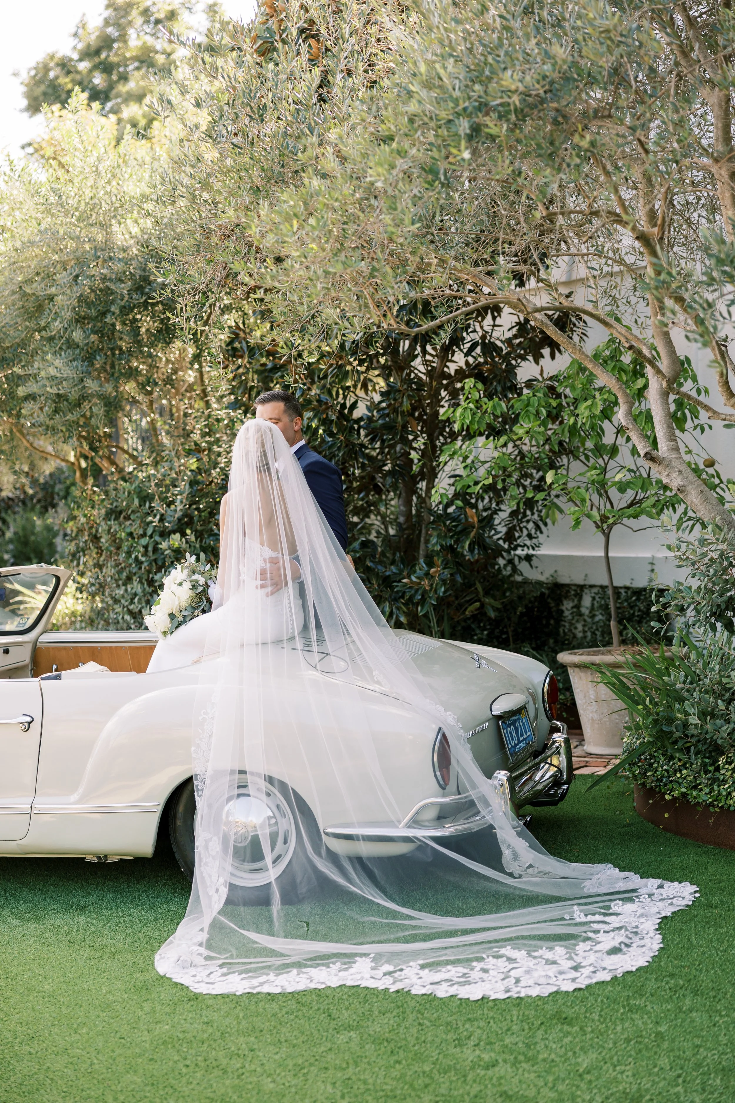Bride and groom kissing on a vintage car outdoors, surrounded by greenery, with the bride wearing a long veil and holding a bouquet at a Pensacola Wedding Venue.