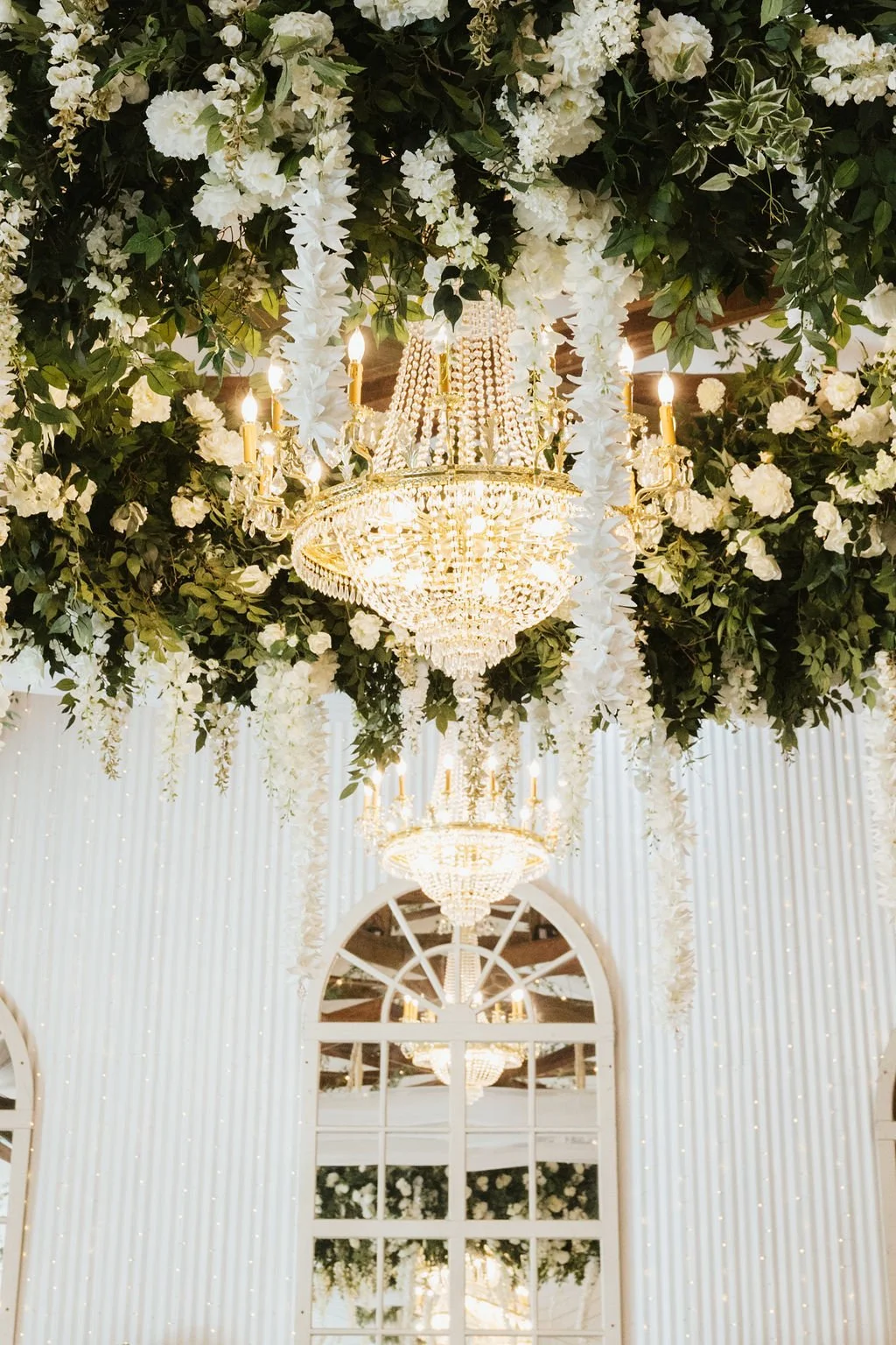 Elegant chandelier hanging from a floral ceiling with white flowers and greenery, reflected in arched windows with white curtains and string lights in a Pensacola Wedding Venue.
