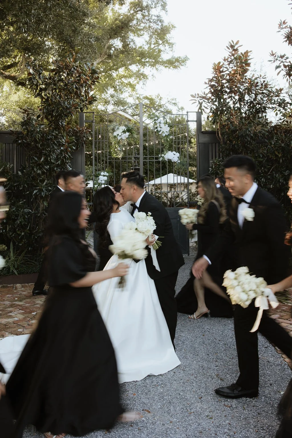 A wedding ceremony with a bride and groom kissing, surrounded by bridesmaids and groomsmen holding bouquets. The scene is outdoors, with trees and a decorative floral and metal arch in the background.