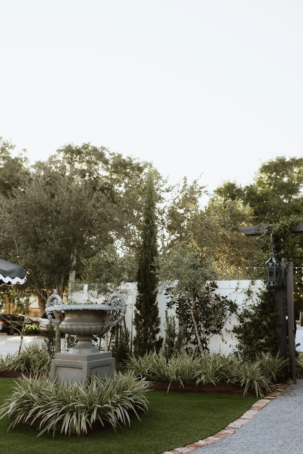 Ornamental garden with a large decorative fountain, lush greenery, trees, and a white fence in the background.