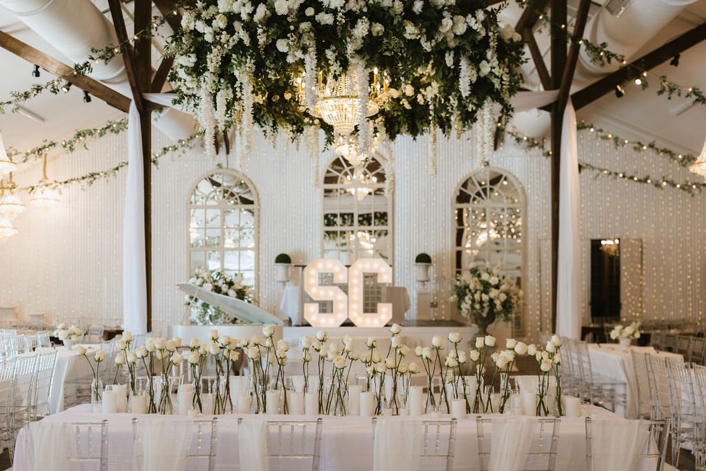 Pensacola Wedding reception decorated with white flowers, candles, and string lights, featuring a backdrop with large illuminated initials 'SC' at the center, and a ceiling adorned with floral arrangements and drapes.
