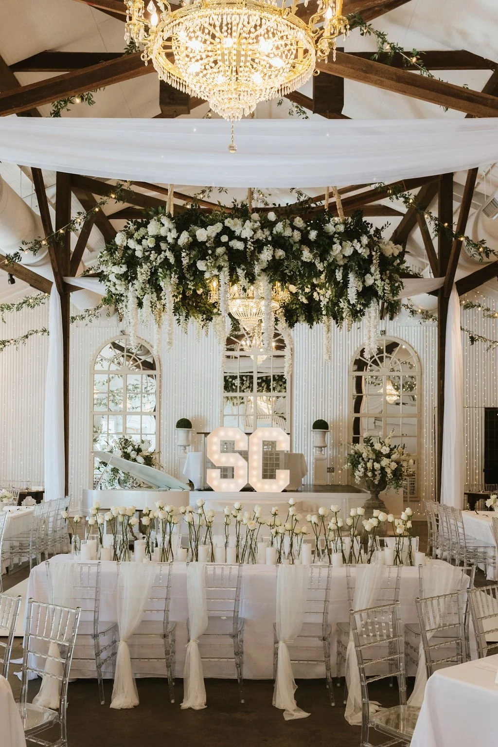 Elegant wedding reception setup with a large floral chandelier, white drapery, and a decorated stage featuring large illuminated "SC" initials, white roses, and greenery.