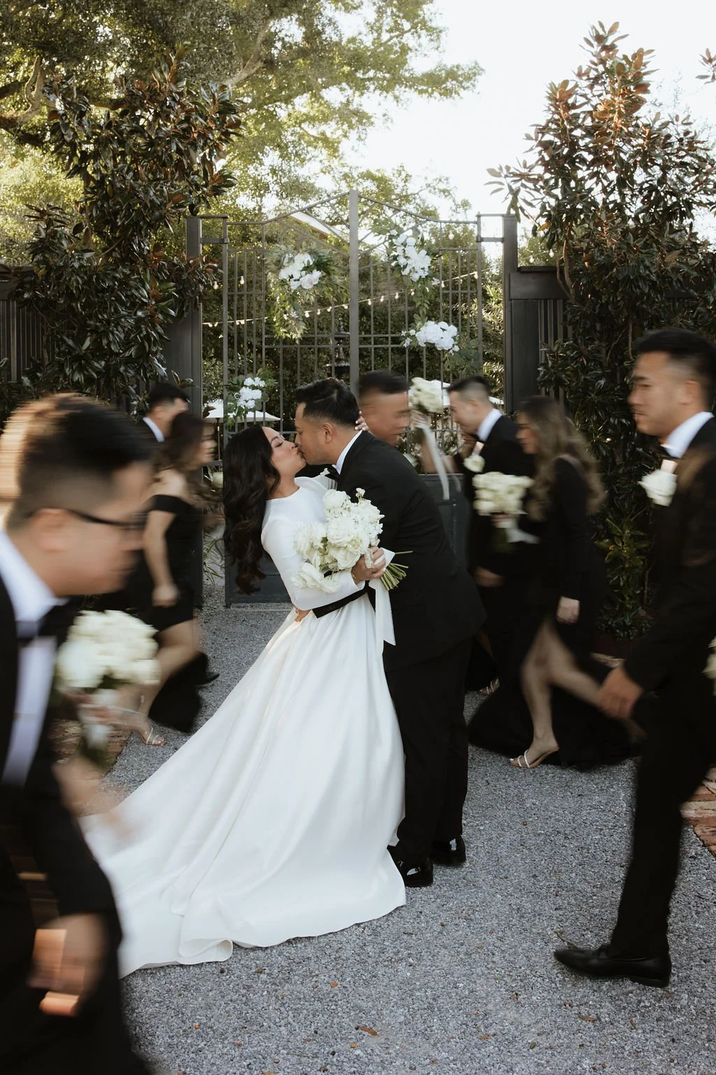 A bride and groom kiss while holding a bouquet of white flowers, surrounded by wedding guests dressed in black suits and dresses, walking under a decorative arch with white flowers at an outdoor wedding reception.