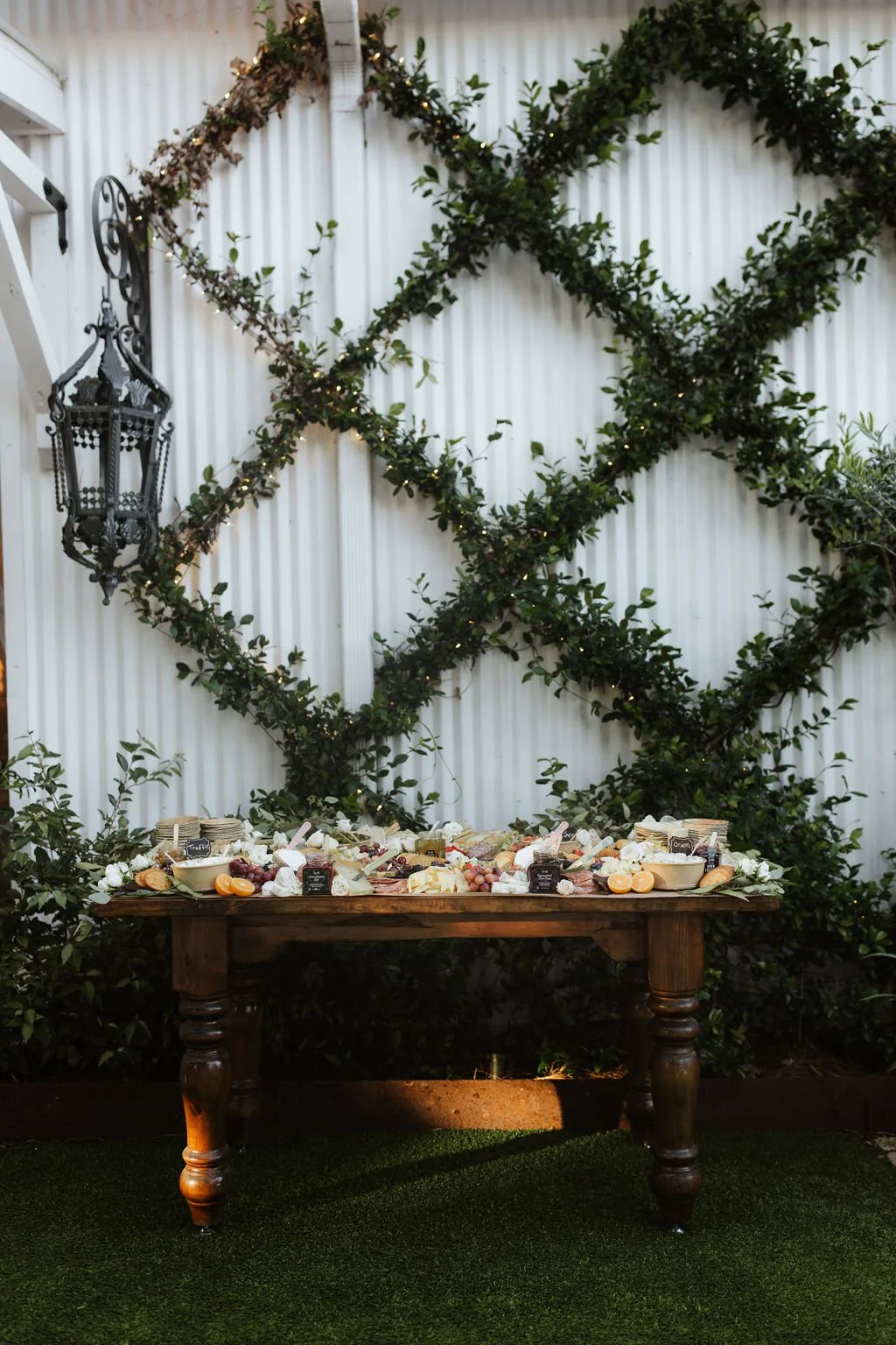 A cheese and charcuterie display on a wooden table set outdoors against a white corrugated metal wall decorated with a large diamond-shaped green vine pattern and string lights. An ornate black lantern hangs on the left side.