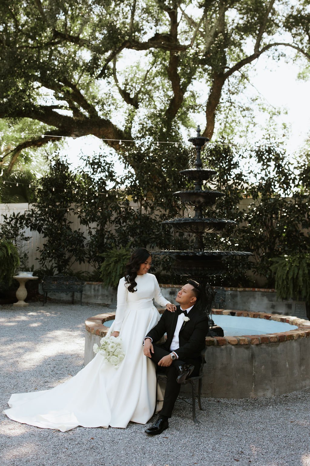 A bride and groom in wedding attire outdoors in front of a fountain, surrounded by trees and greenery, with the bride holding a bouquet of white flowers.