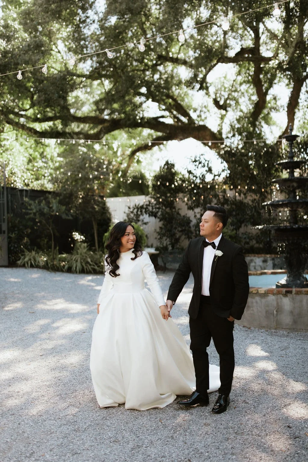 A bride and groom holding hands and walking outdoors, with the bride smiling and the groom looking at her, under a large tree with string lights hanging above, in a Pensacola garden setting.