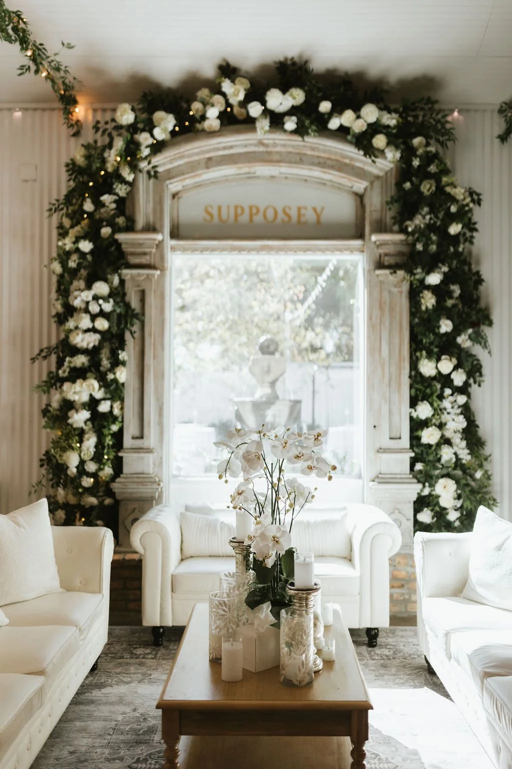 A cozy living room decorated with white floral arrangements and greenery, featuring a vintage fireplace with the sign 'Supposey' above it, white sofas, a wooden coffee table with candles and orchids, and a large window allowing natural light.