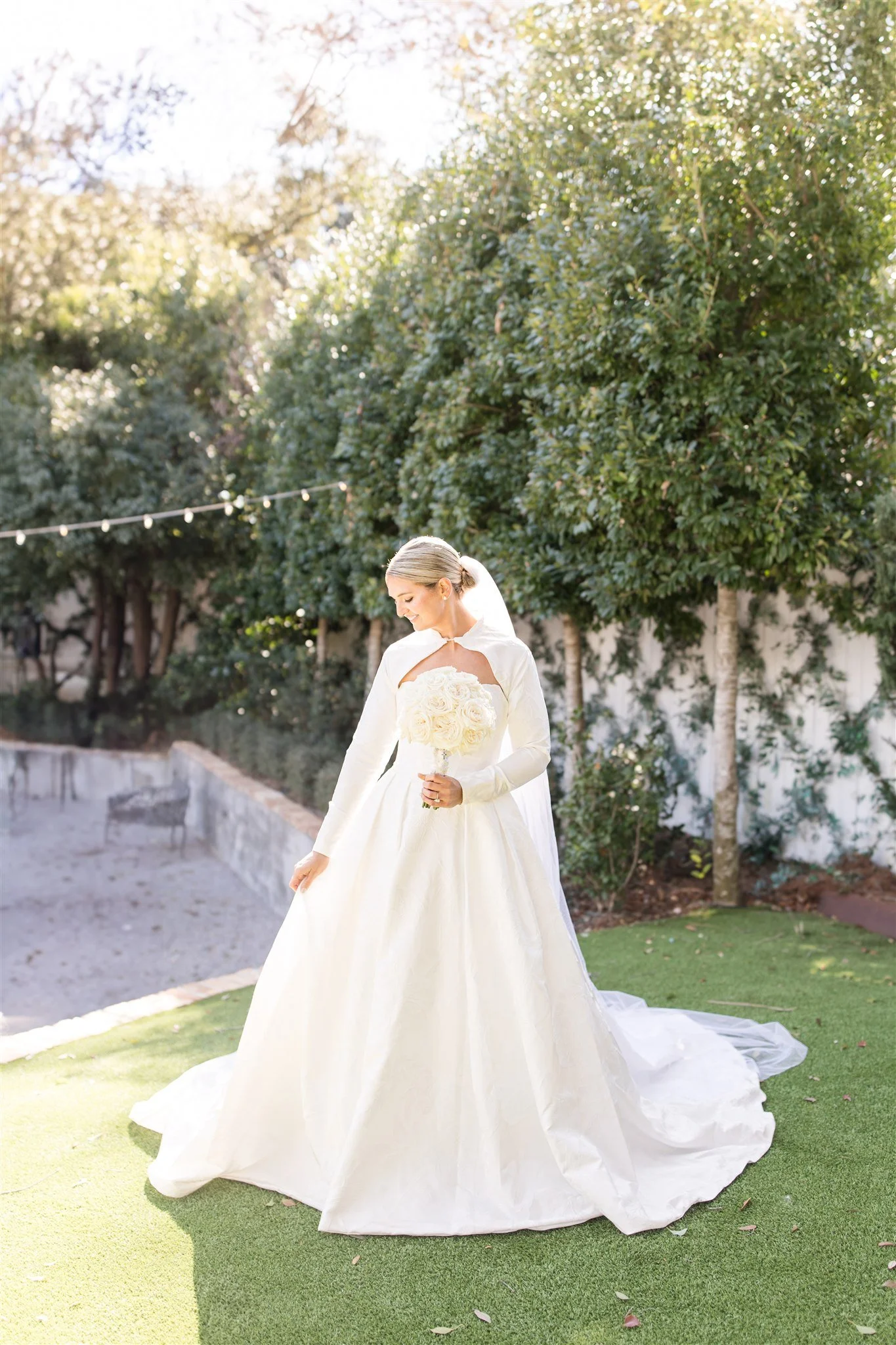 A bride in a white wedding dress holding a bouquet of white roses in an outdoor garden setting with trees and string lights