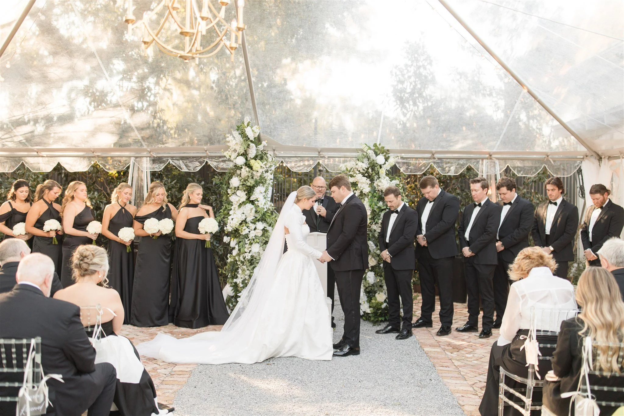 A wedding ceremony taking place under a clear tent with the bride and groom holding hands and kneeling, surrounded by bridesmaids in black dresses and groomsmen in black tuxedos, with guests seated on either side and floral arrangements in the backgr