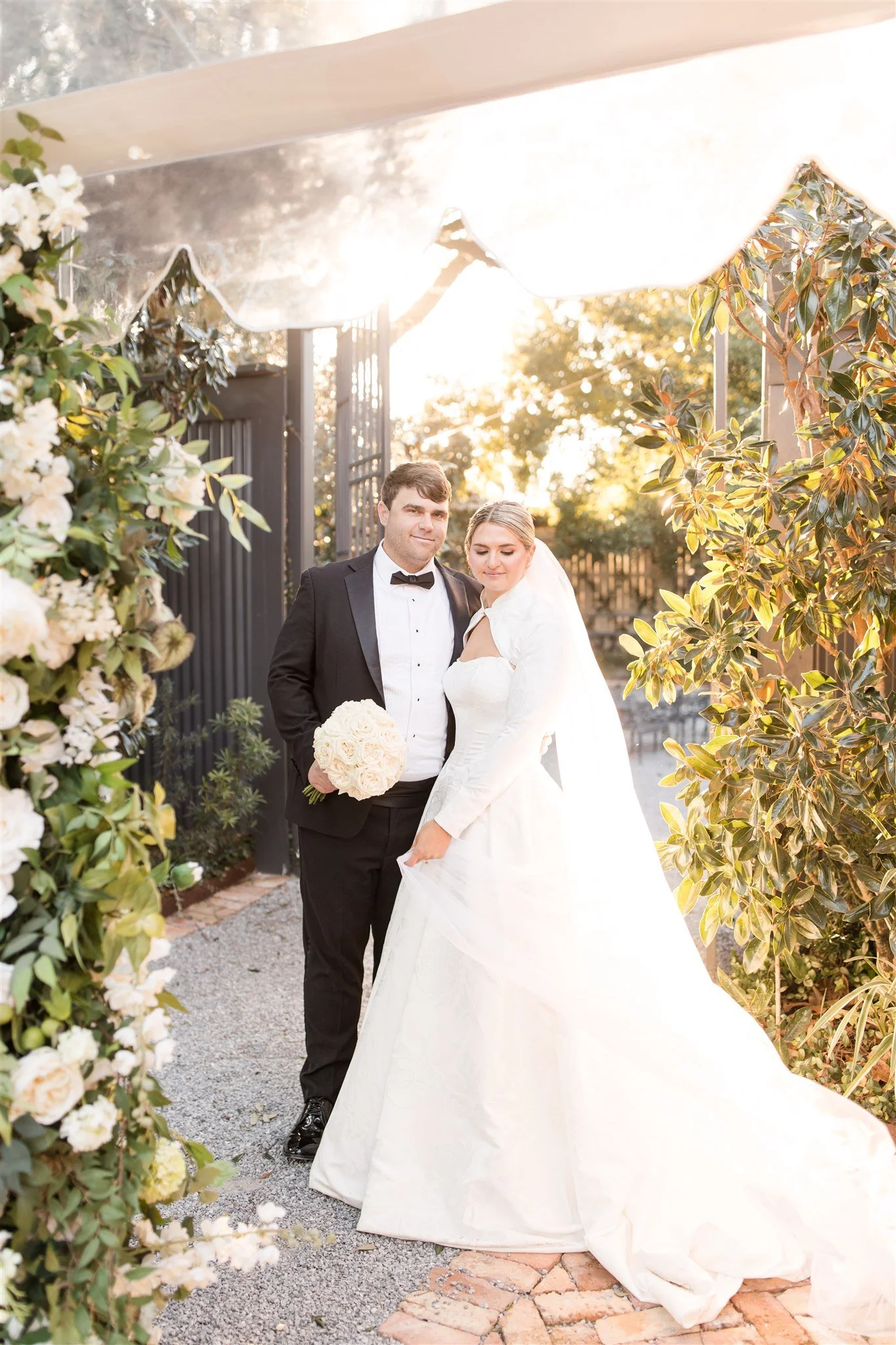 A bride and groom standing together outdoors during sunset, surrounded by greenery and floral decorations, with the bride holding a bouquet of white roses.