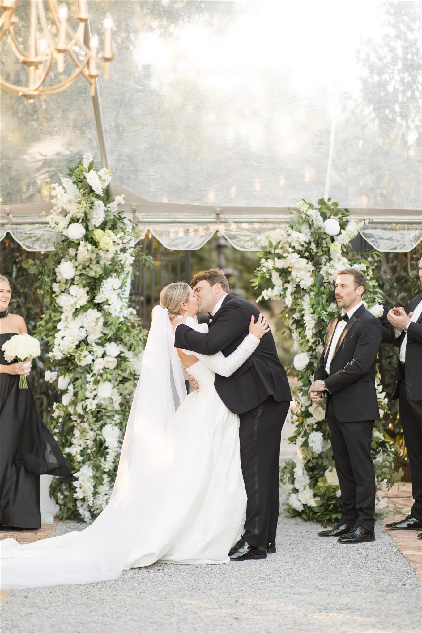 A bride and groom share a kiss during their wedding ceremony, surrounded by flowers and witnesses at a Pensacola Wedding Venue.