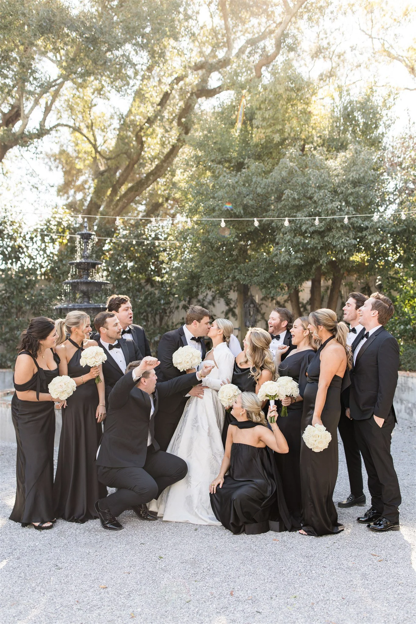 A wedding party of thirteen people, including the bride and groom, are gathered outdoors on a gravel surface with trees and string lights in the background. The bride is in a white gown, and the groom and others wear black tuxedos. Bridesmaids wear b