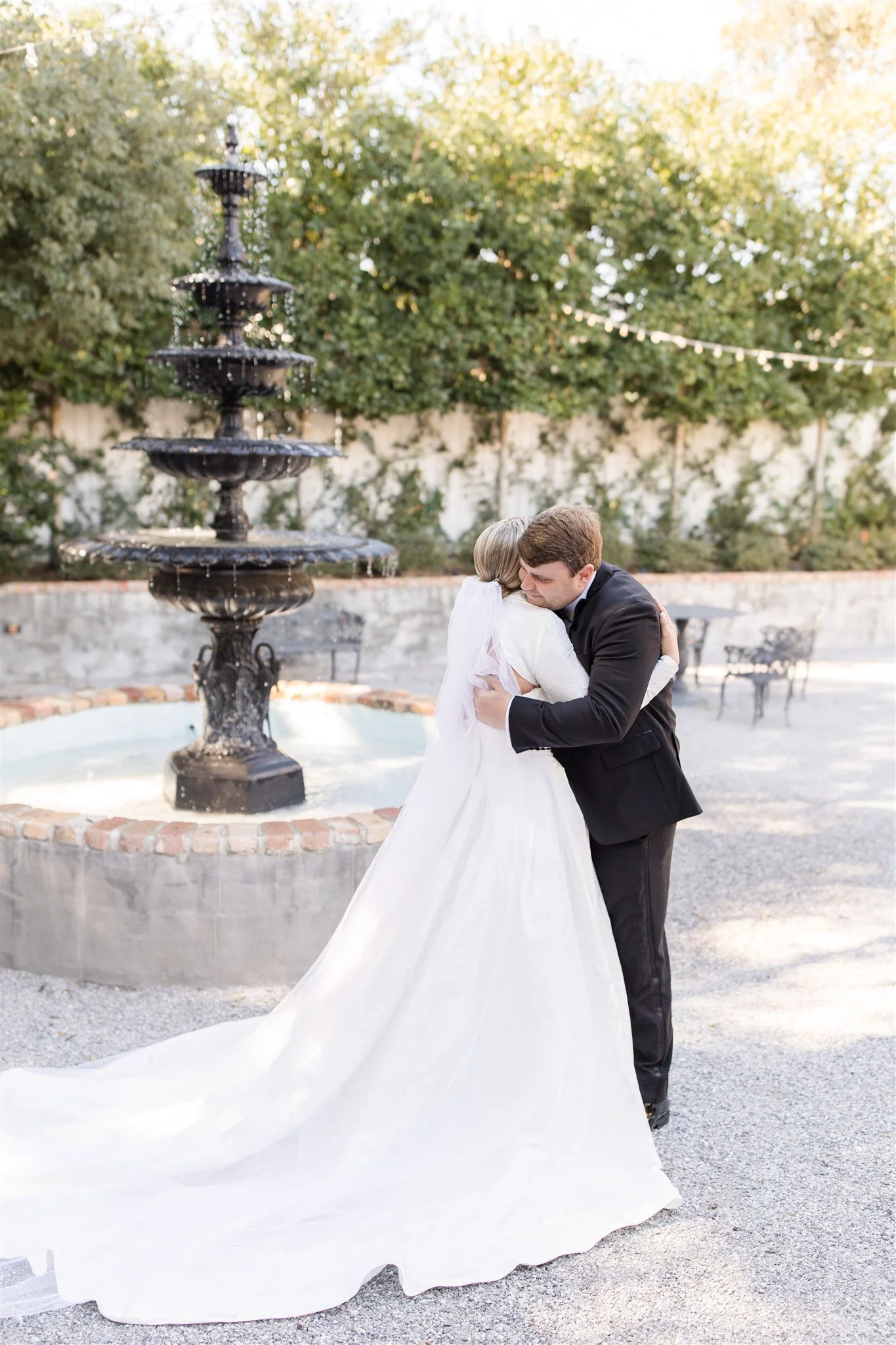 A bride and groom hug in front of a fountain outdoors during wedding.