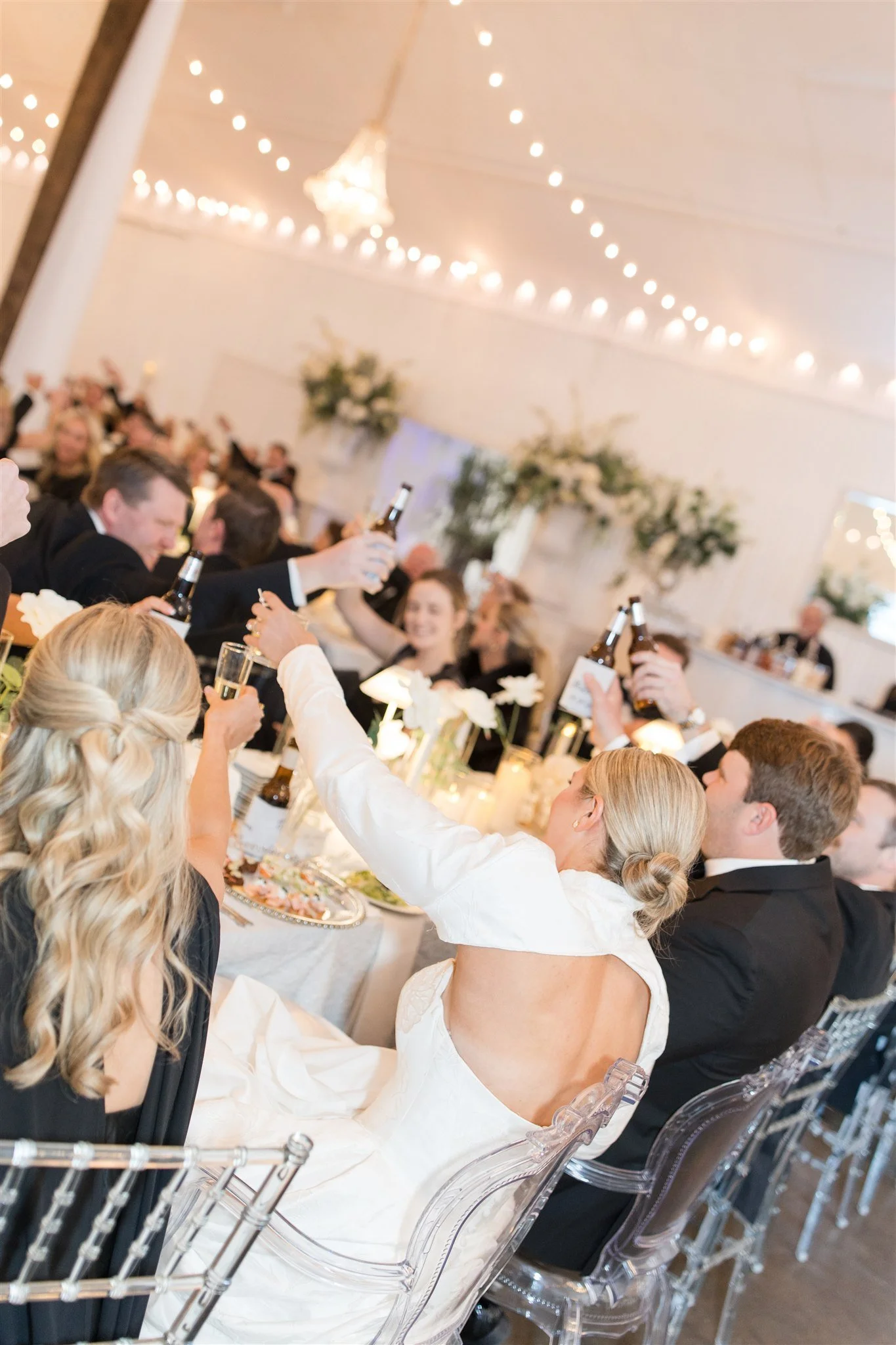 People at a wedding reception raising glasses in a toast, with elegant decor, flowers, and string lights in the background.
