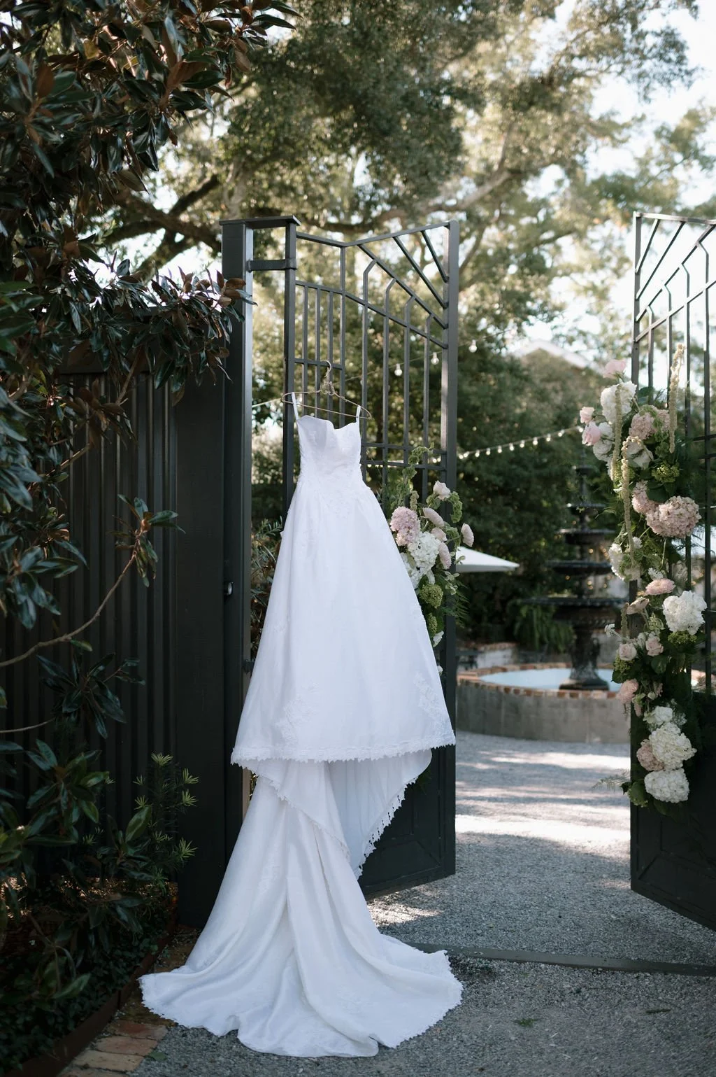 A white wedding dress hanging on a gate adorned with flowers, with trees and an outdoor fountain in the background.