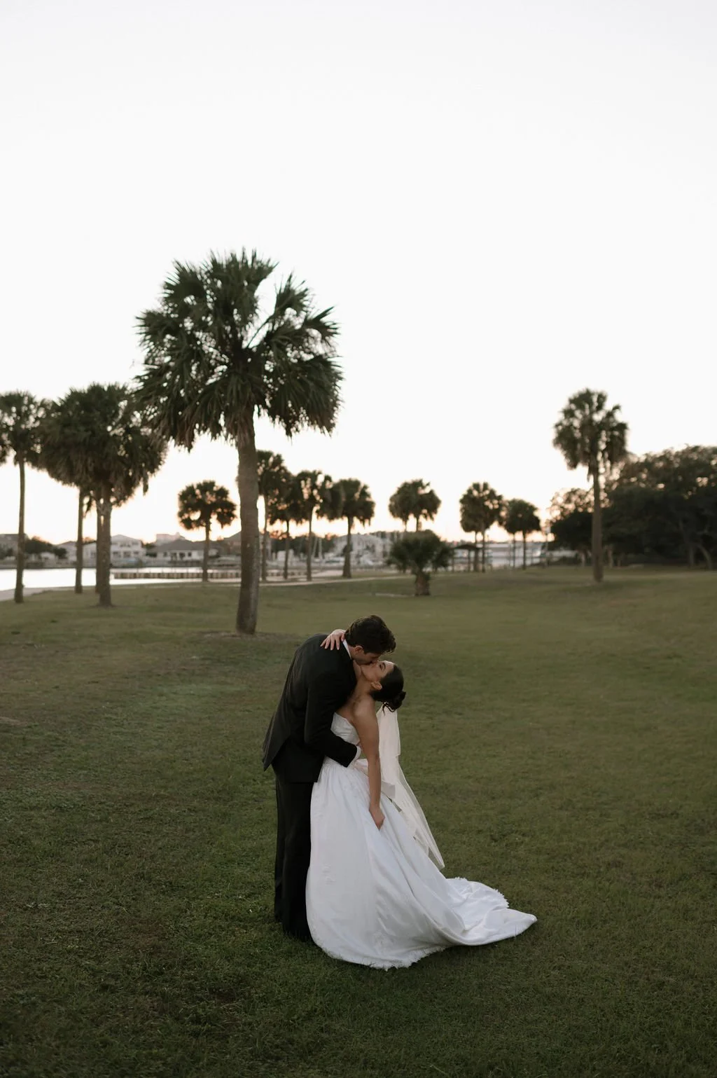 A couple in wedding attire sharing a kiss outdoors on a grassy field with palm trees and water in the background during sunset.