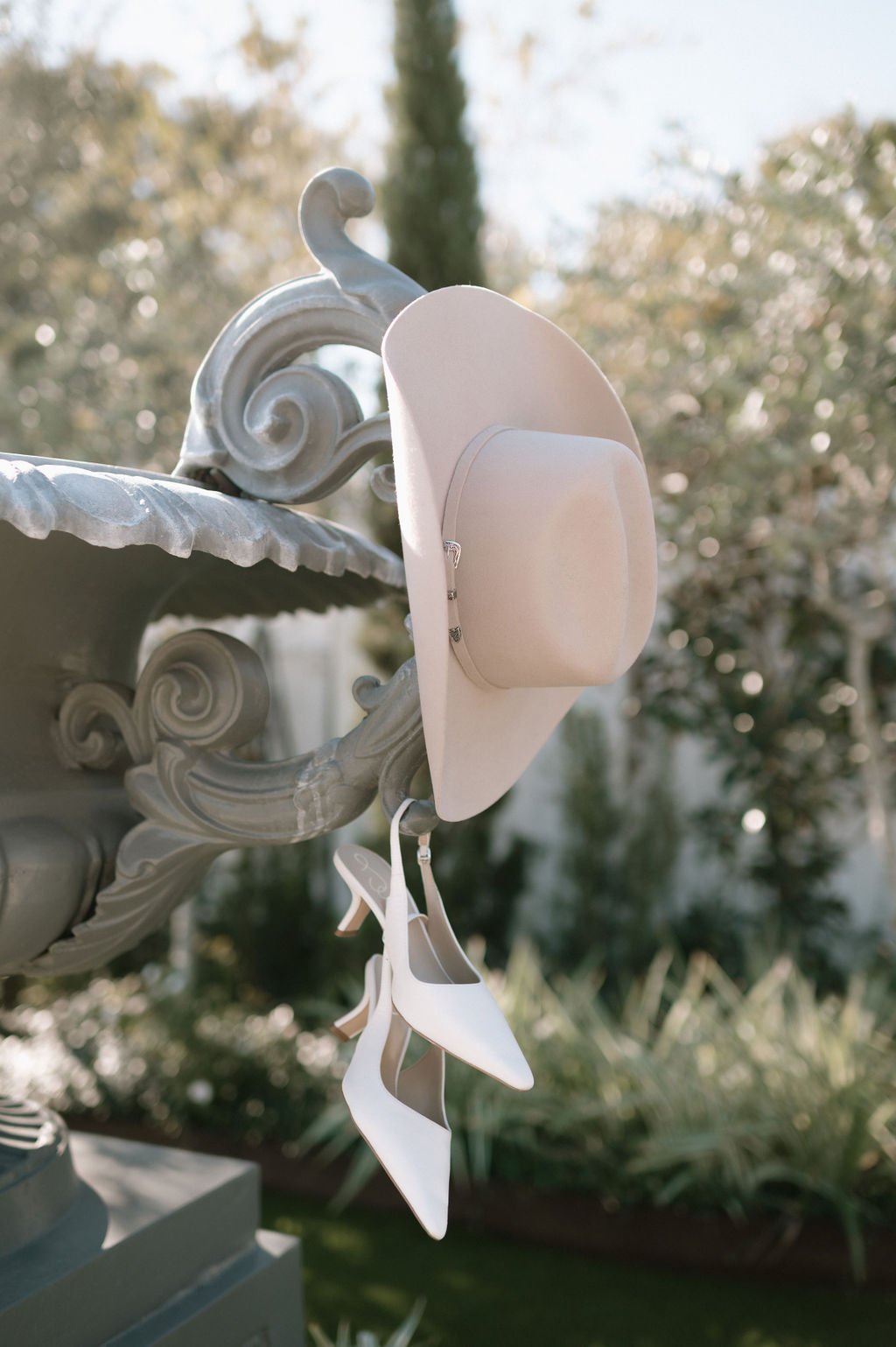 A white hat, white shoes, and a pair of shiny white high heels are hanging on the decorative corner of a vintage-style, ornate metal fountain, with a blurred background of trees and greenery.