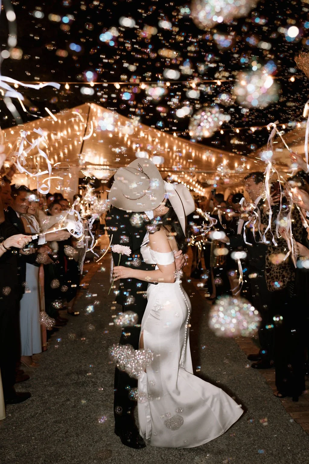 A bride and groom kissing under bubbles and string lights at their wedding reception.