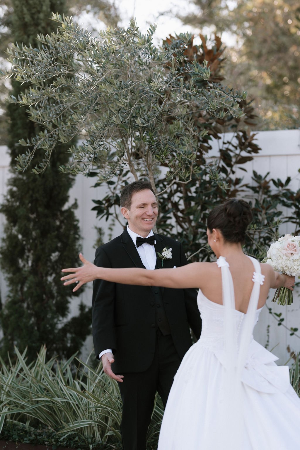 A newly married couple, a man in a black tuxedo and bow tie and a woman in a white wedding dress, greeting each other outdoors in a garden with trees and plants.