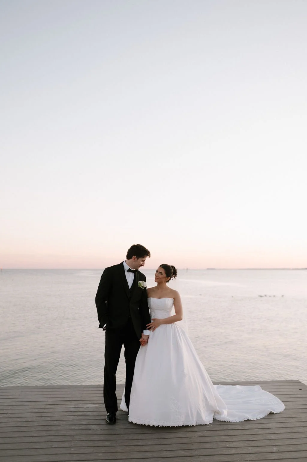 A bride and groom in wedding attire standing on a wooden dock by the water at sunset, holding hands and smiling at each other.