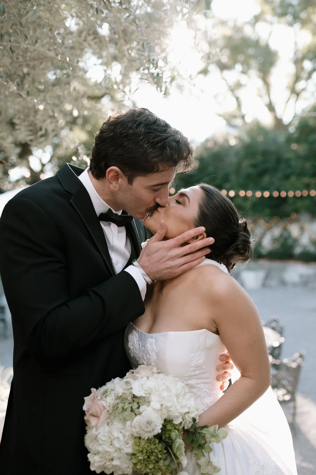 A newlywed couple sharing a kiss at their wedding outdoors, with the groom in a black tuxedo and the bride in a strapless white wedding gown holding a bouquet of flowers.