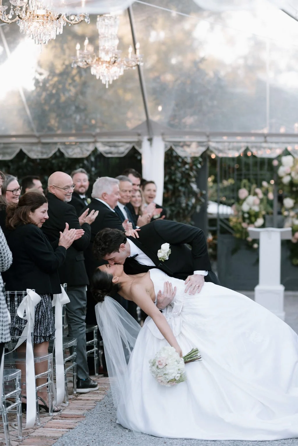 A bride and groom sharing a kiss at their wedding ceremony, with guests clapping and smiling in the background under a decorated canopy.