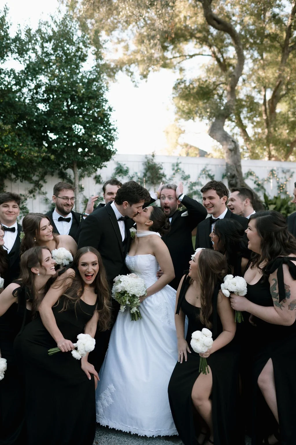 A wedding party of diverse group of people celebrating outdoors, with a bride and groom in the center, surrounded by bridesmaids and groomsmen, all dressed elegantly.