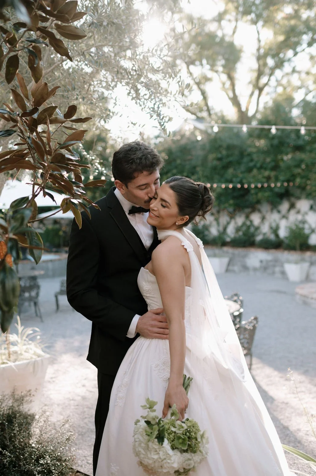 A bride and groom embrace outdoors at sunset, with trees and string lights in the background. The bride is holding a bouquet of white and green flowers, and the groom is kissing her forehead.