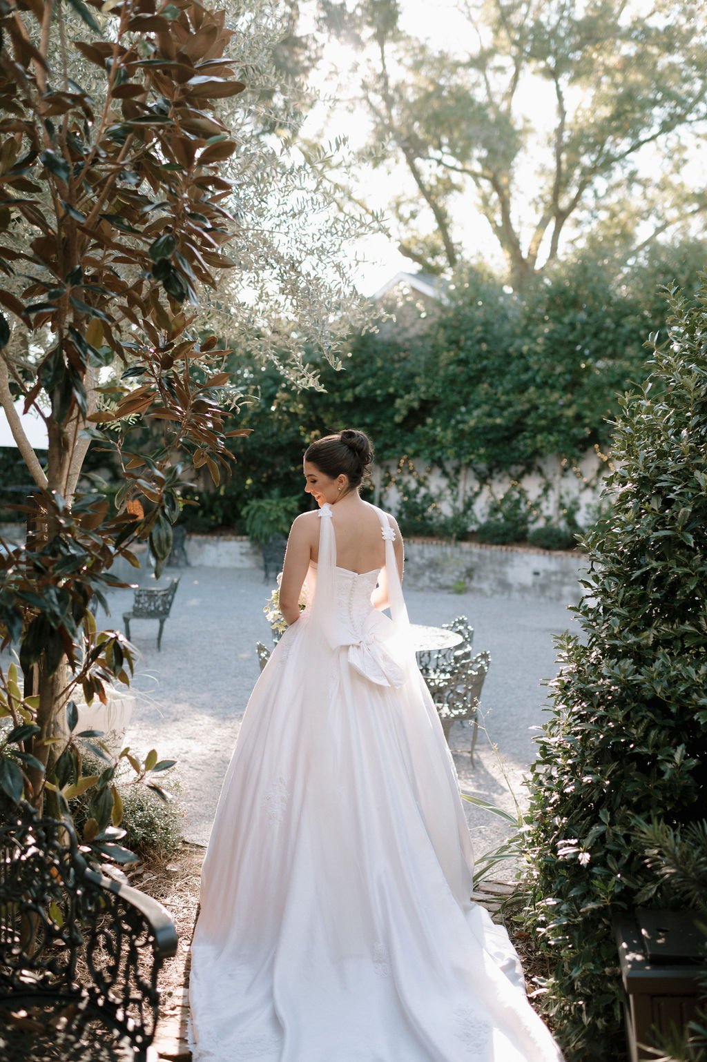 A bride in a white wedding gown with a large bow on the back stands in a garden with greenery and outdoor furniture, illuminated by sunlight.