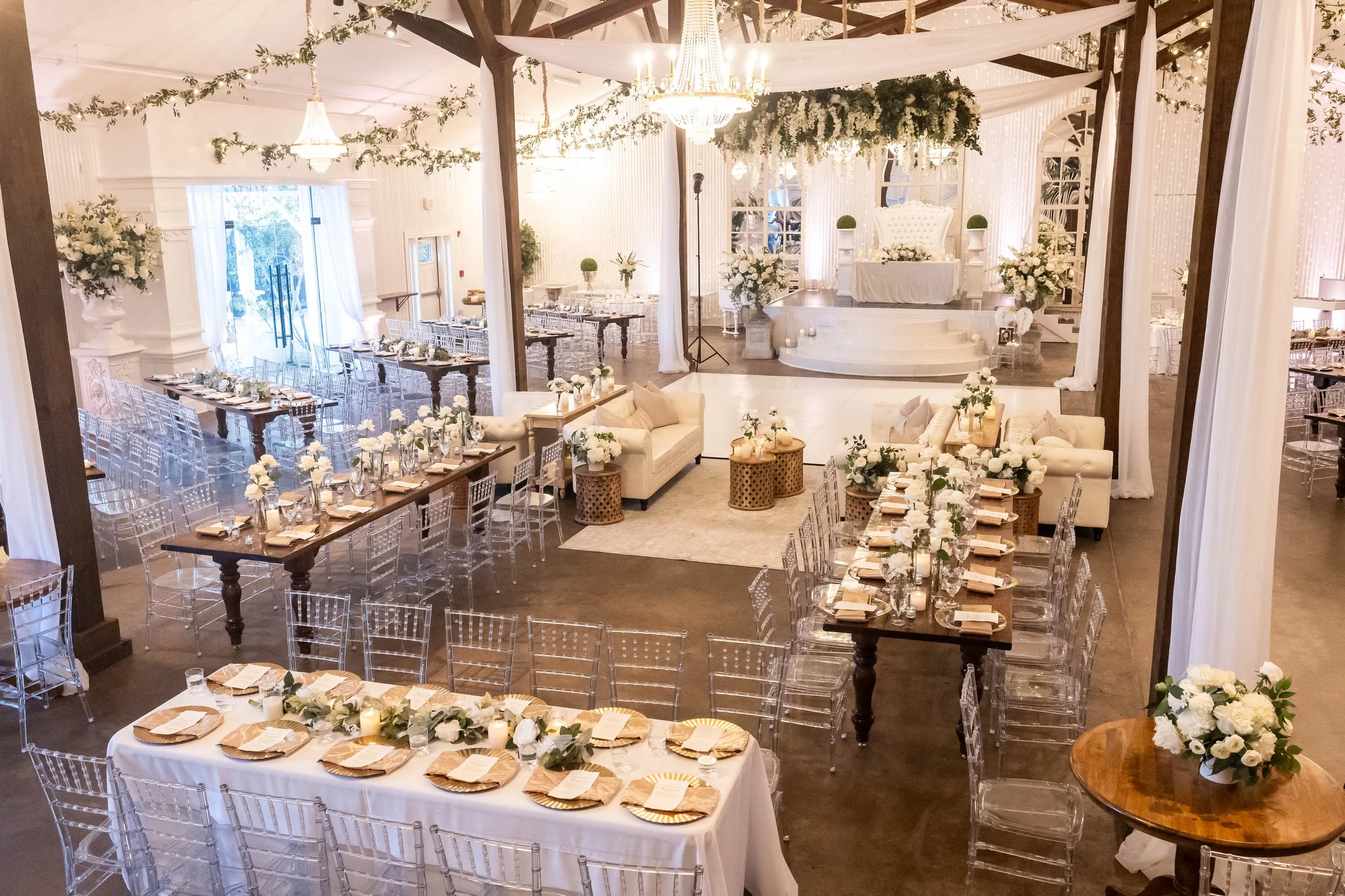 Elegant Pensacola wedding reception setup with long dining tables, floral centerpieces, and a stage with a throne-style chair decorated with white flowers, chandeliers, draped curtains, and white and neutral-colored decor.