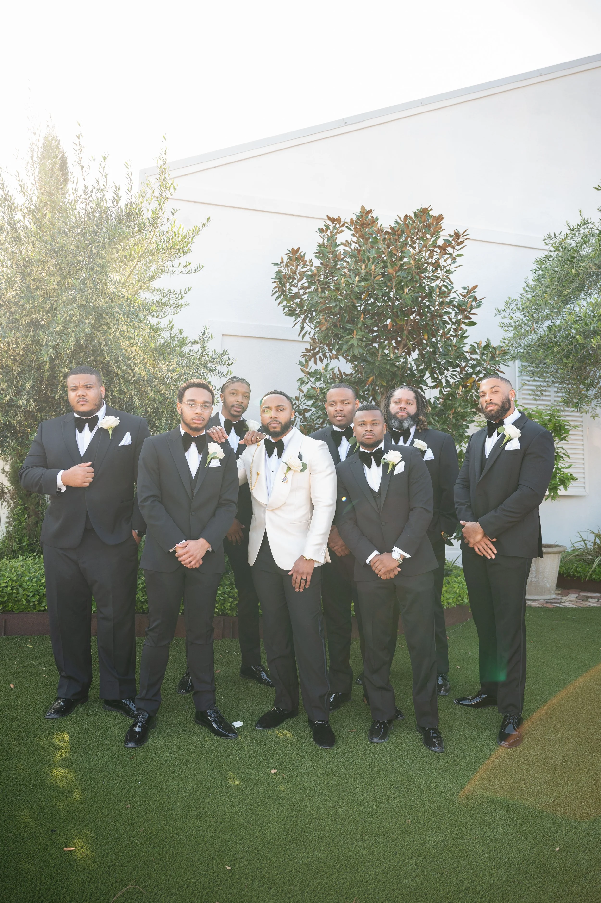 A group of nine men in tuxedos, with one in a white jacket, pose outside on a green lawn in front of bushes and trees.
